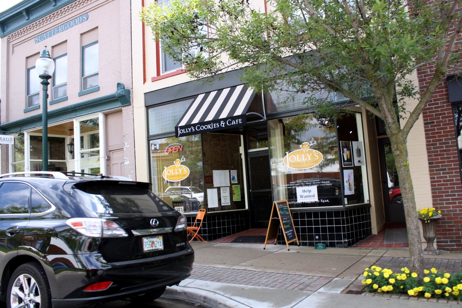 A row of downtown storefronts with a striped awning, a cafe sign in the window and yellow flowers along the curb.