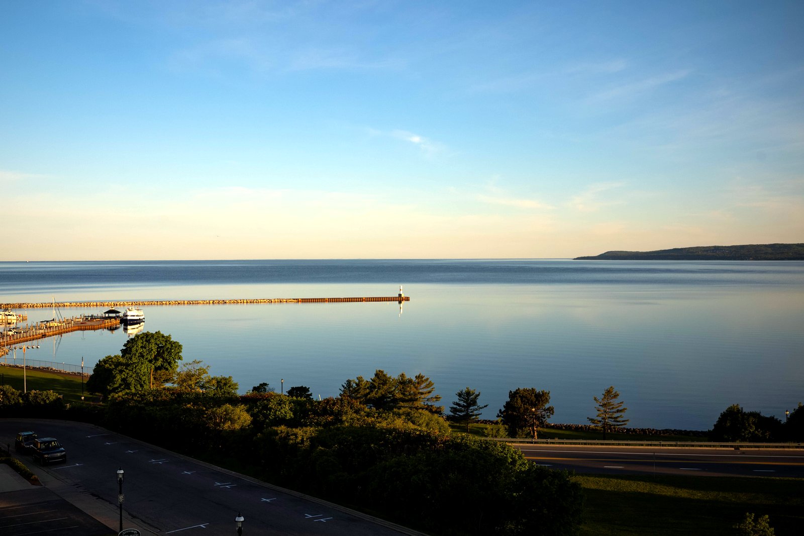 Calm blue water of Little Traverse Bay under a clear evening sky, with a breakwall and small marina at left and trees and a road in the foreground.