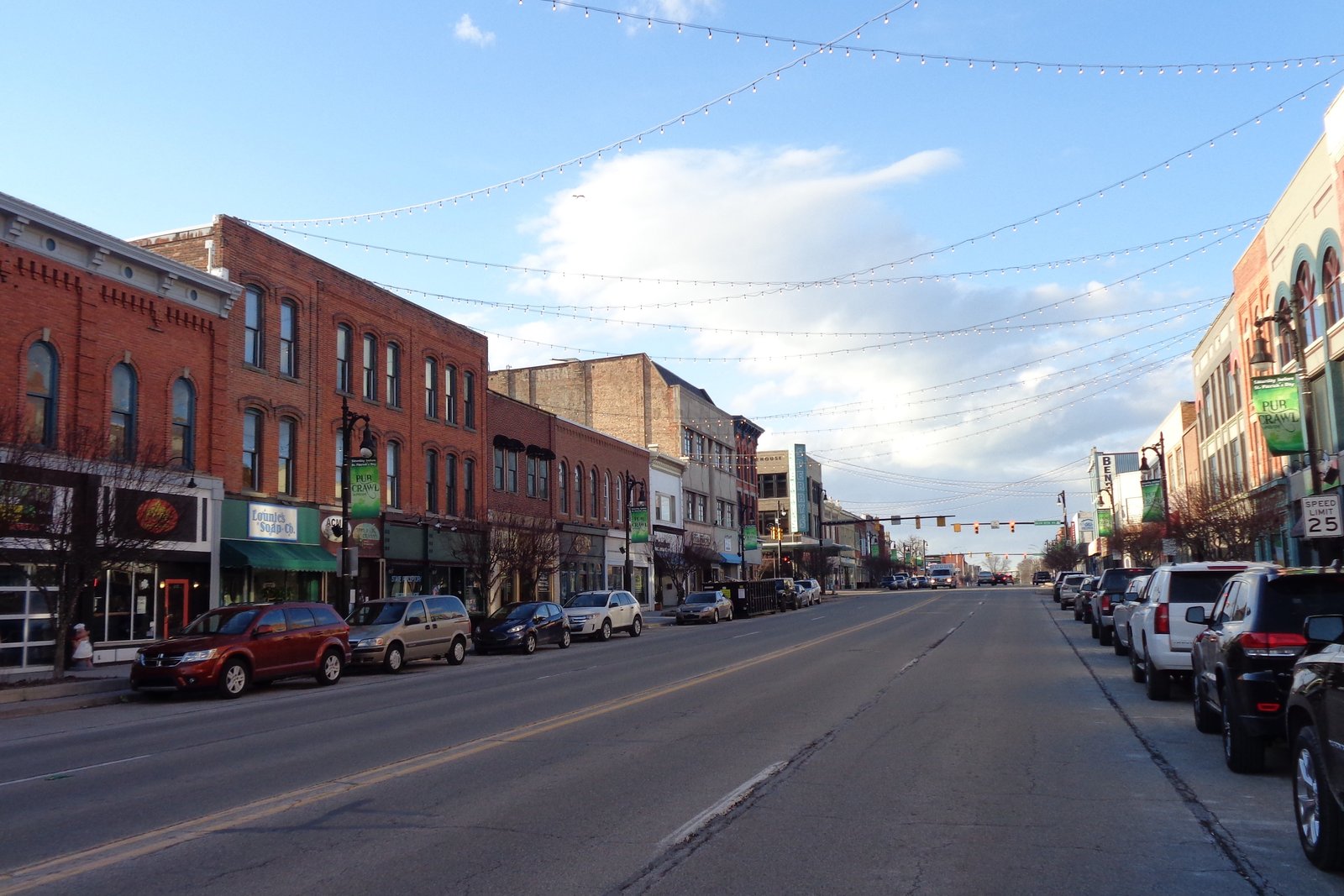 A downtown Port Huron street lined with two- and three-story brick storefronts, parked cars and strings of lights overhead under a blue sky.