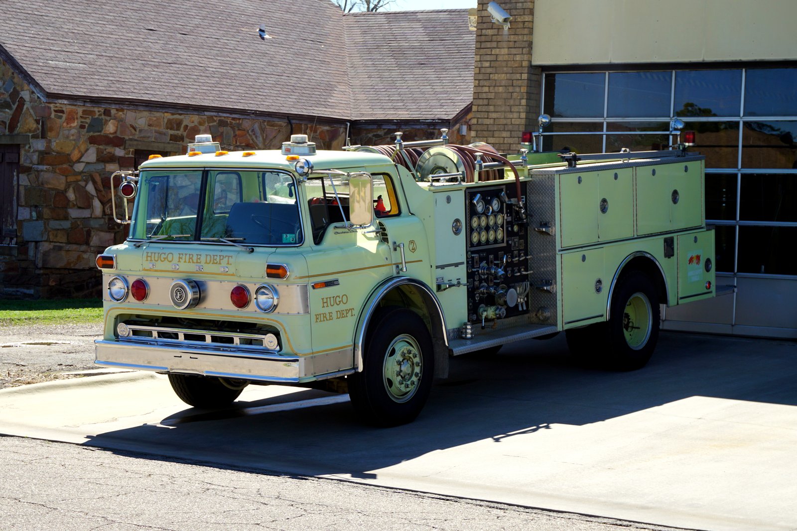 A red fire engine labeled Engine 2 sits parked outdoors beside a building, with trees and a grassy lawn behind it.