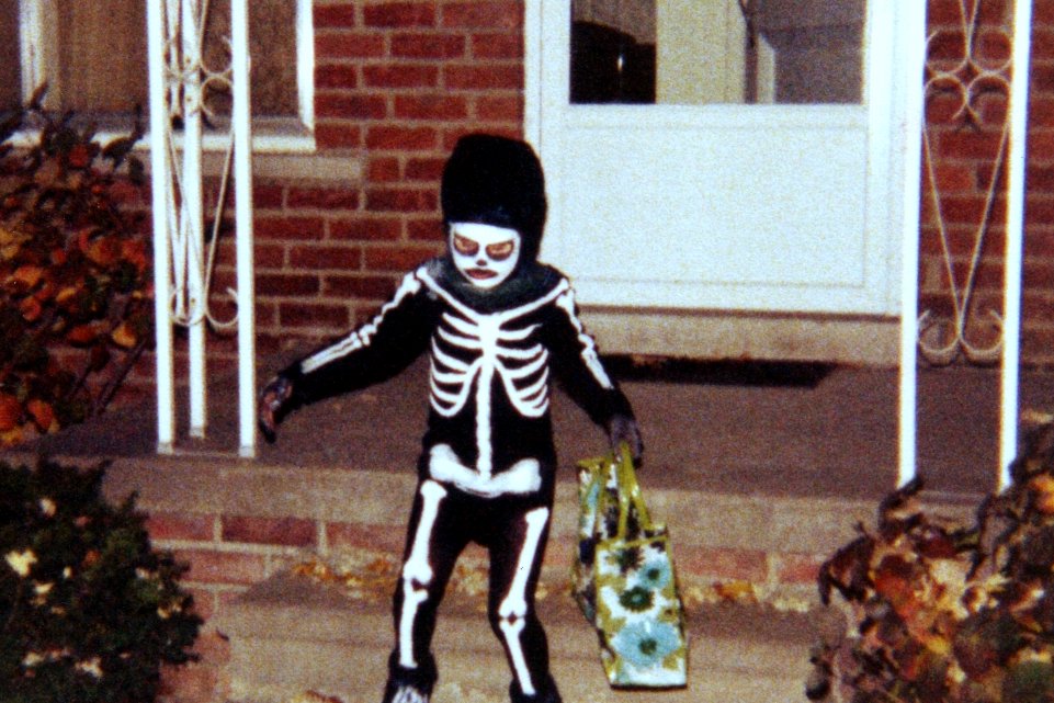 A child in a skeleton costume and mask holds a treat bag on the steps of a brick house strewn with fallen leaves.