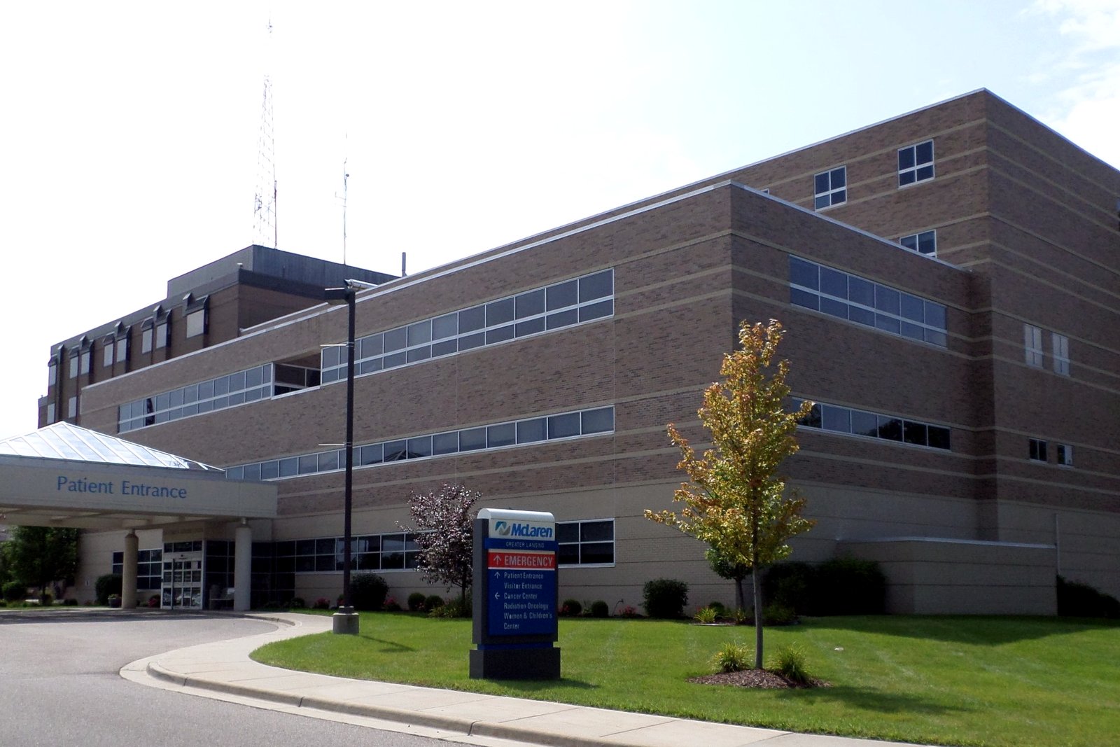 A tan brick hospital building with a covered patient entrance and a McLaren emergency sign on a landscaped lawn under a hazy sky.