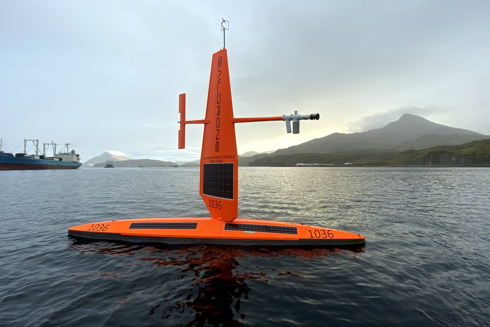 An orange uncrewed sailboat with a tall rigid wing sail floats on gray water, with a cargo ship and mountains in the distance.