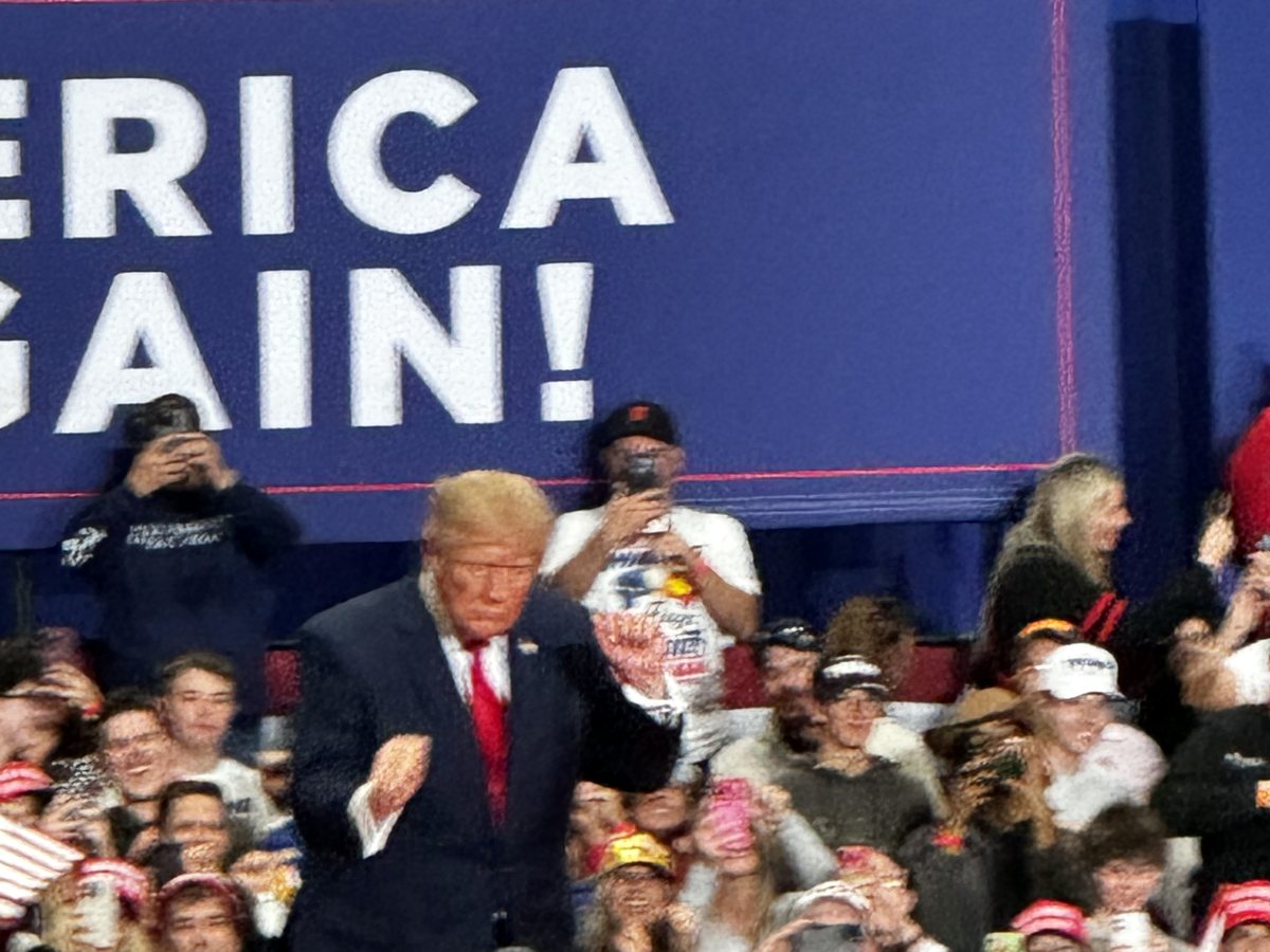 President Donald Trump walks along the front of the stage past cheering supporters, many holding up phones, under a large Make America Great Again banner.