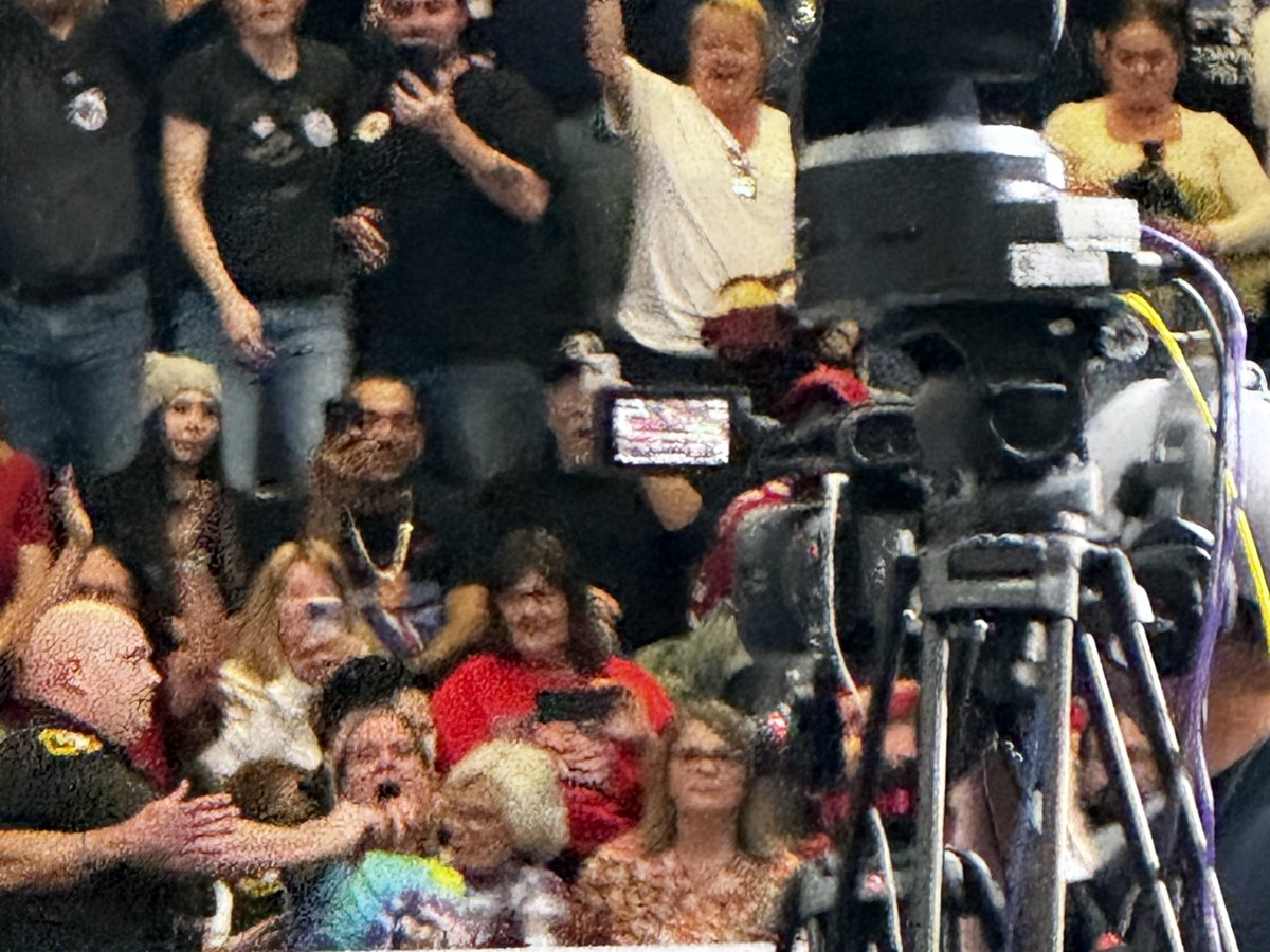 A packed section of rallygoers cheers and records on phones behind a television camera on a tripod inside McMorran Place Arena.