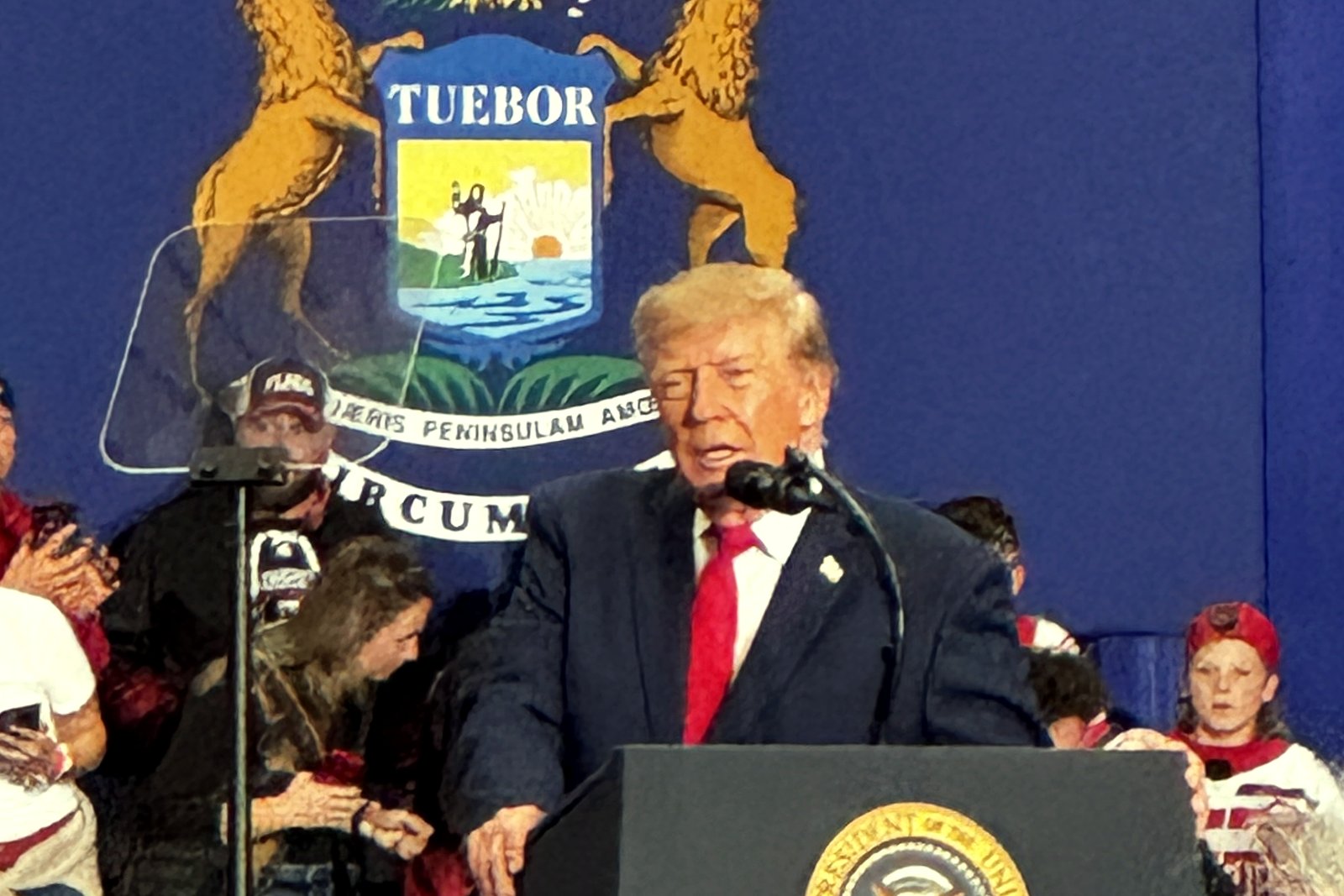 President Donald Trump, in a navy suit and red tie, speaks at a podium bearing the presidential seal in front of a large Michigan state seal, with supporters standing behind him.