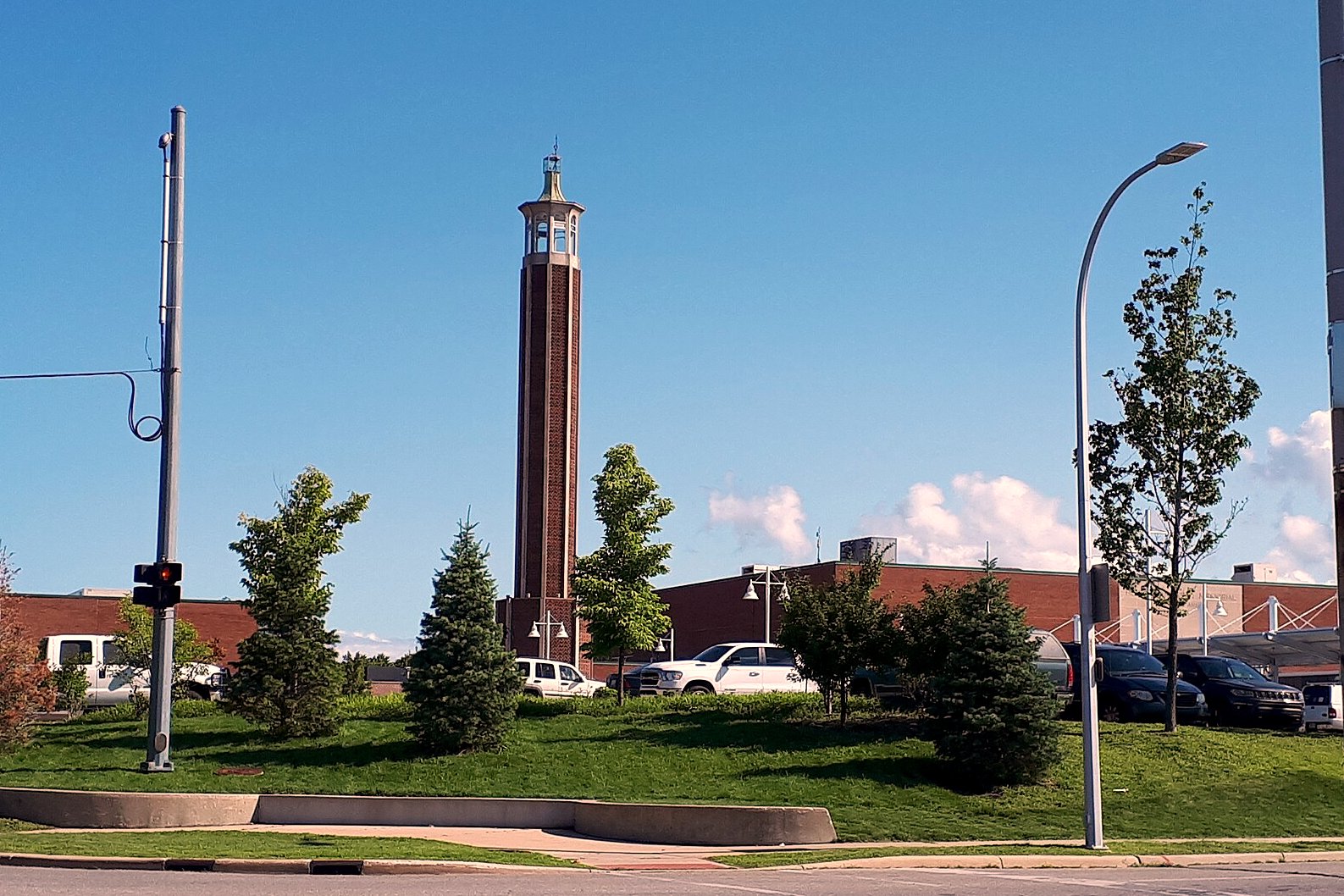A tall brick clock tower rises above trees and a parking lot along a street under a blue sky.
