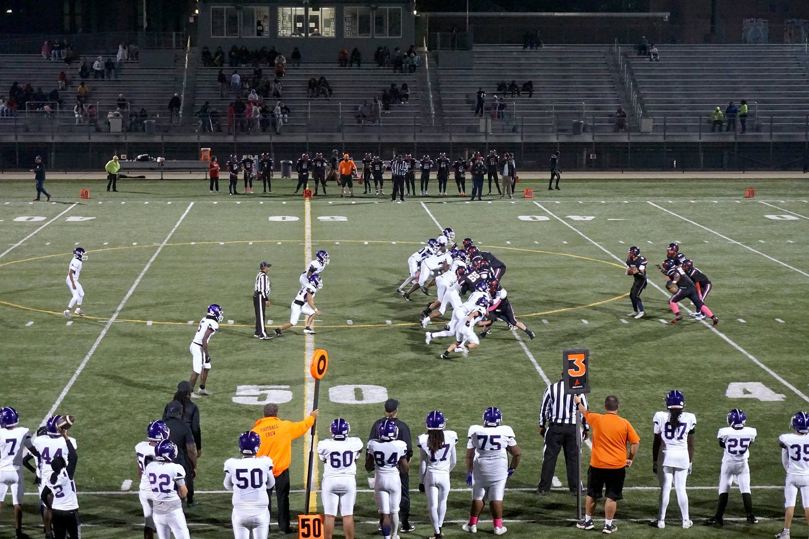 Two high school football teams line up at midfield on a turf field, with players in white and purple and an official on the near sideline.