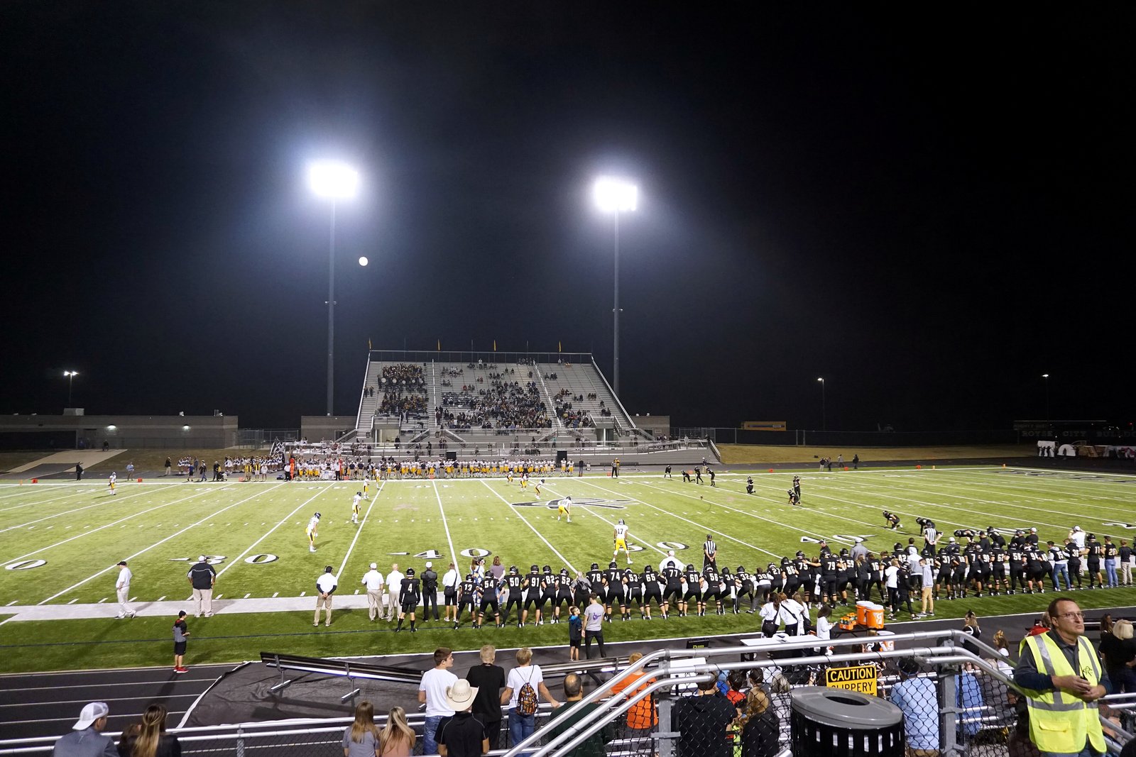 A high school football game kicks off at night under bright stadium lights, with a crowded grandstand across the field and spectators along the near sideline.