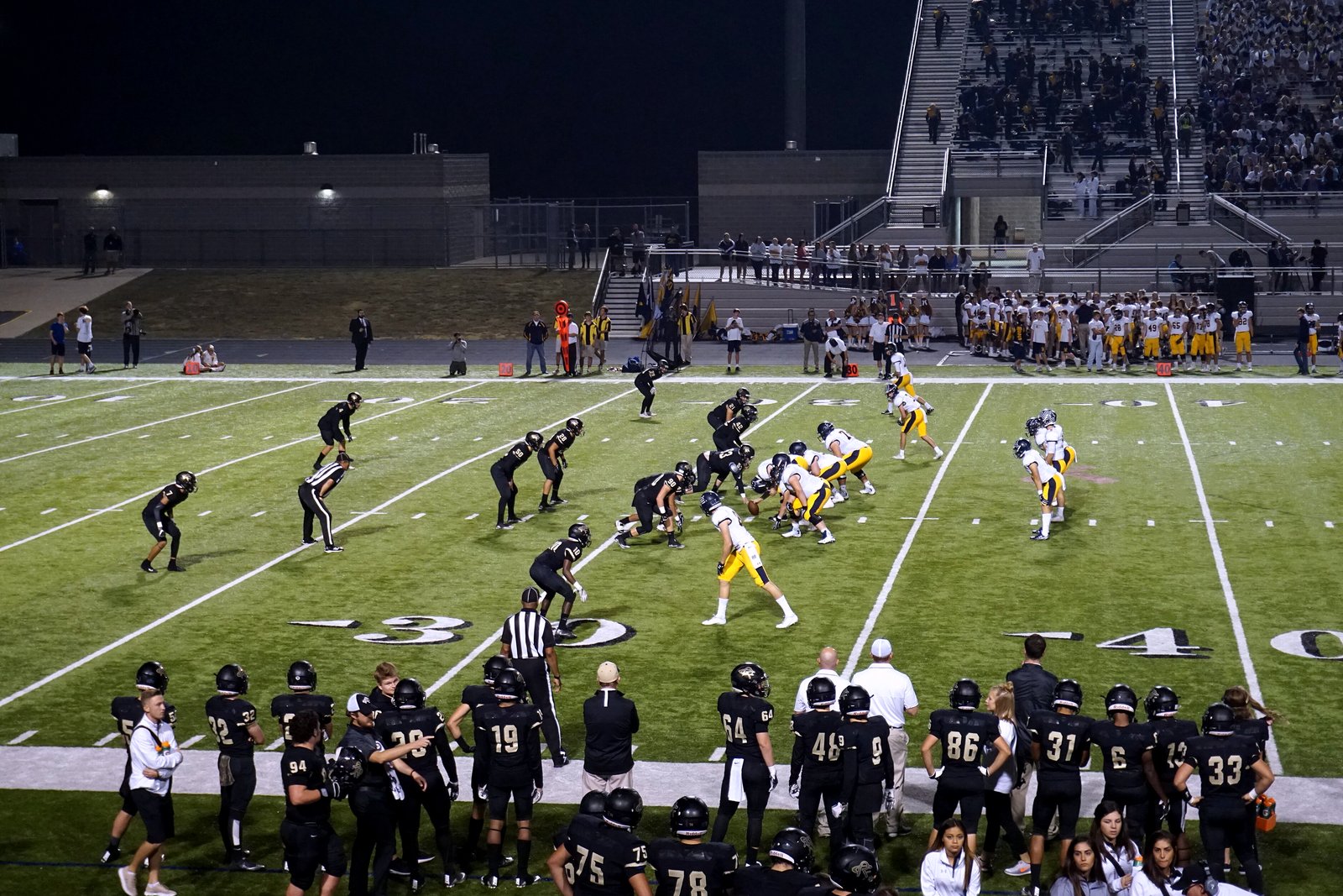 High school football players lined up at the line of scrimmage under stadium lights, a team in black on the near sideline and fans in the stands behind the far sideline.