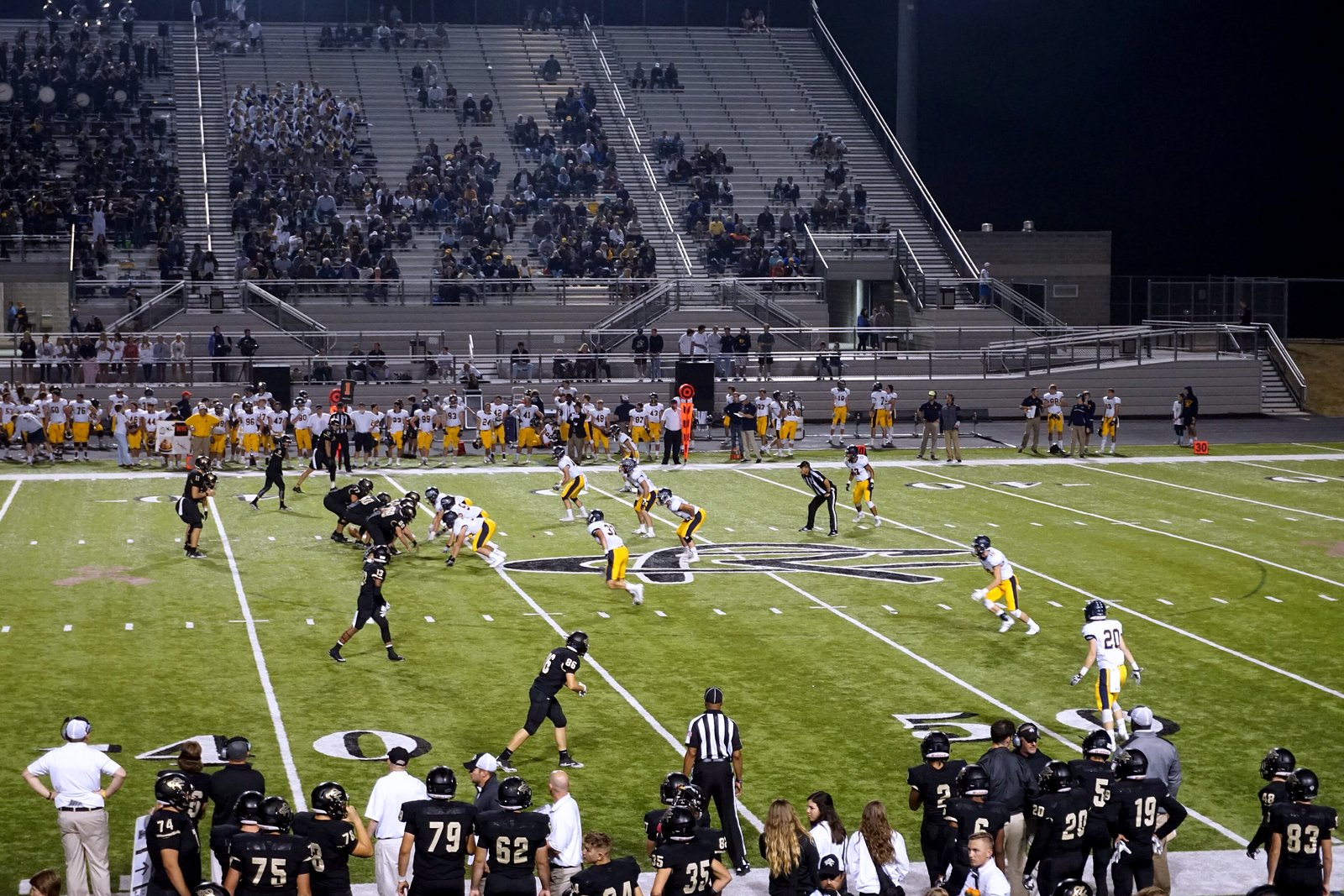 A high school football game at night, with a team in black jerseys lined up against a team in white jerseys and gold pants in front of a crowded grandstand.