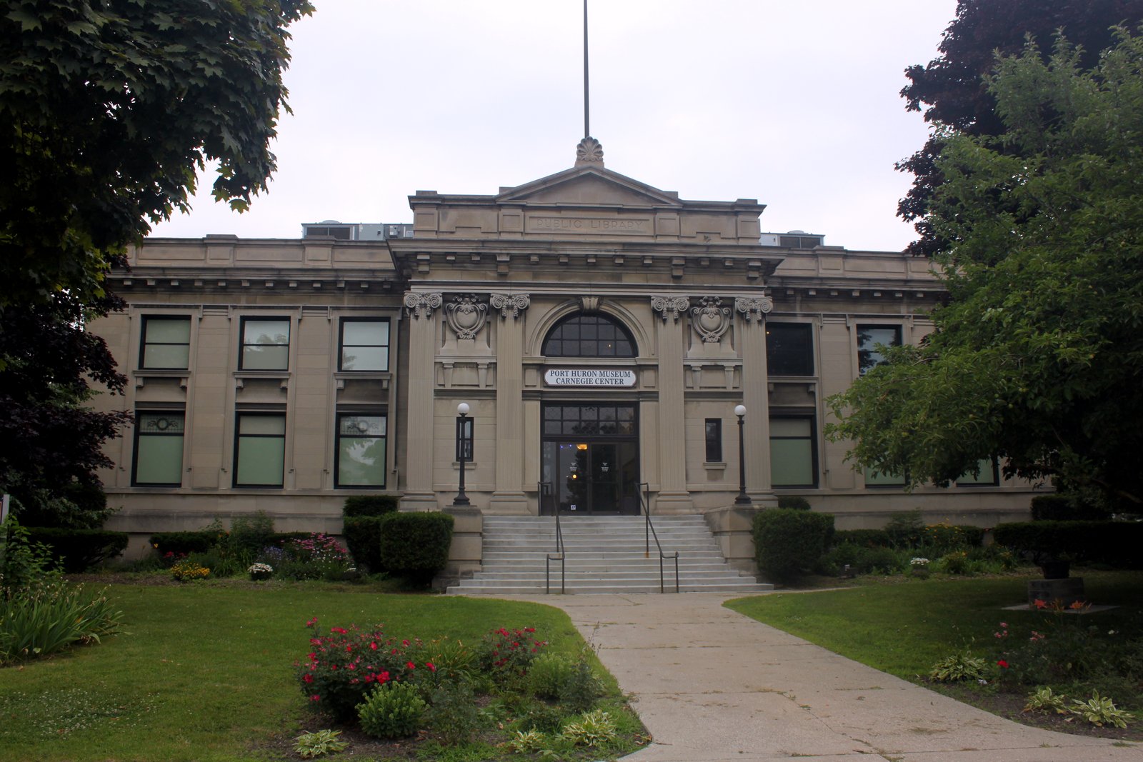 A two-story stone museum building with columns and an arched window, behind a lawn, flower beds and a walkway.