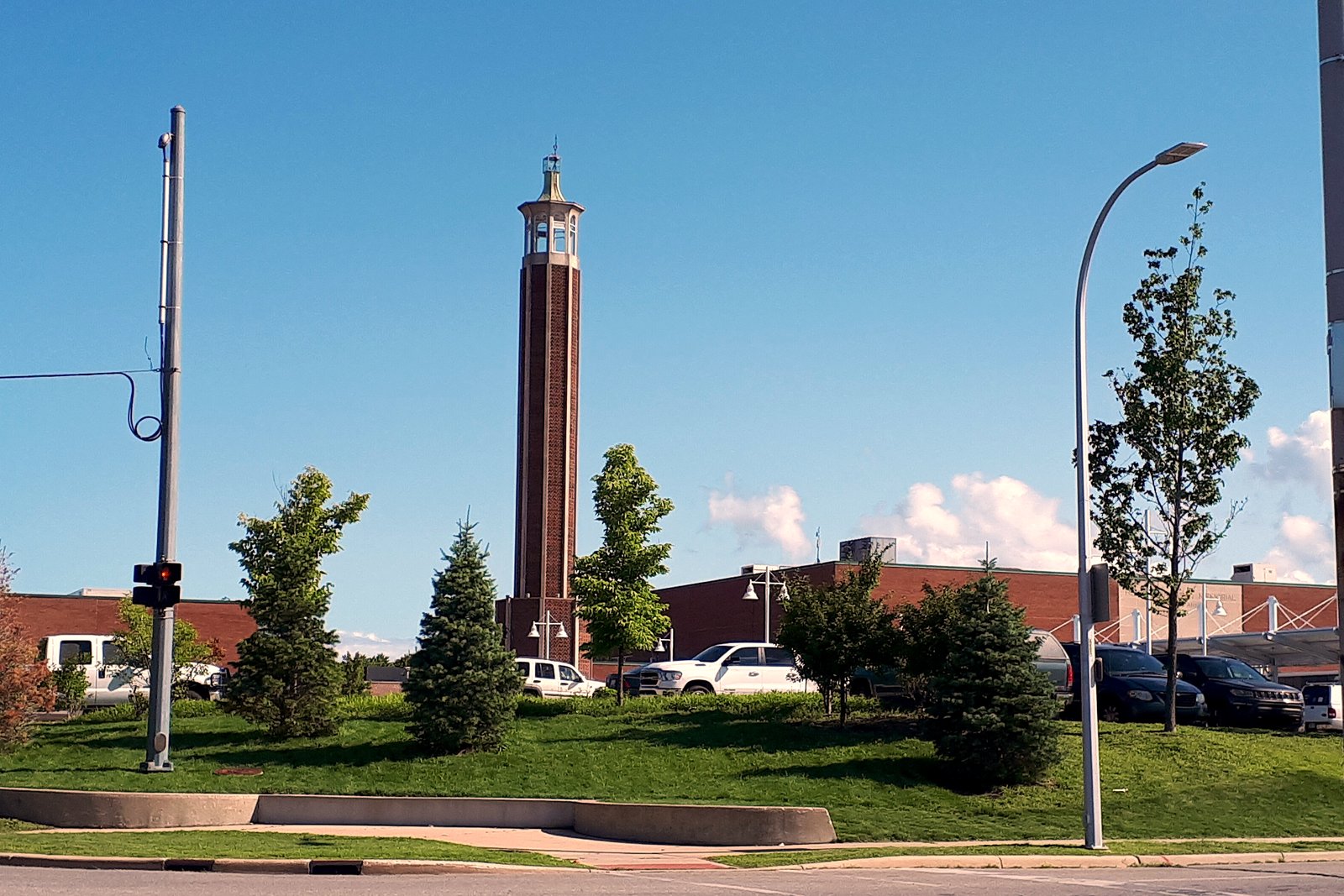 The tall brick tower of the McMorran Place complex rising above a grassy berm and young evergreens, with the arena building and a parking lot behind it under a blue sky.