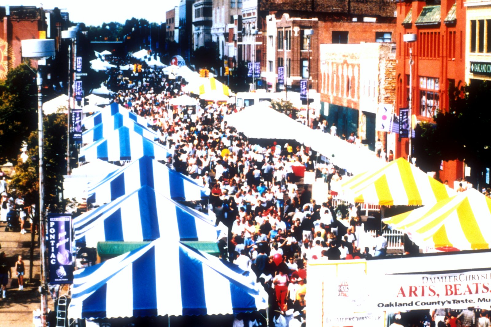 Crowds fill a downtown street lined with blue-and-white and yellow-and-white striped tents during the Arts, Beats and Eats festival, with brick storefronts on both sides.