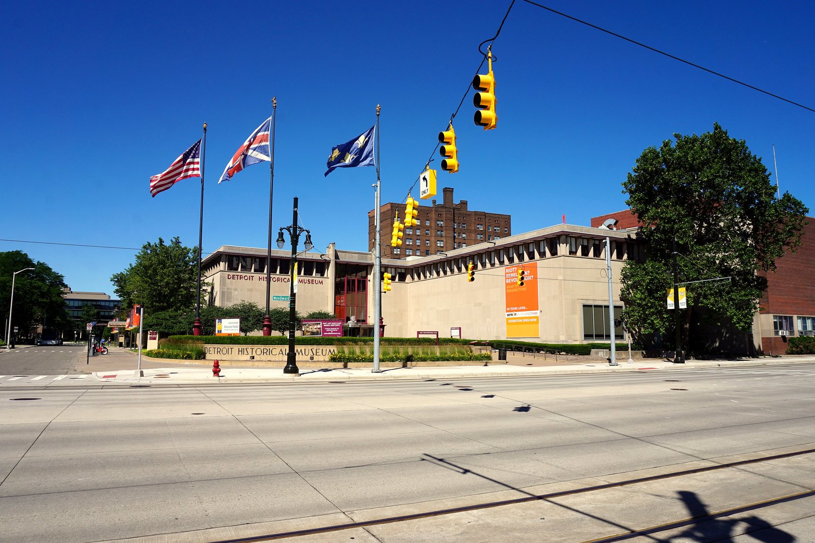 A low stone museum building with flagpoles flying several flags, seen across a wide, empty street on a sunny day.