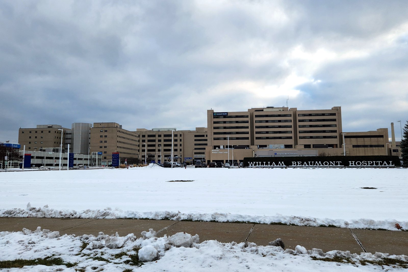 A long tan multistory hospital complex seen across a wide, snow-covered field under a cloudy sky.