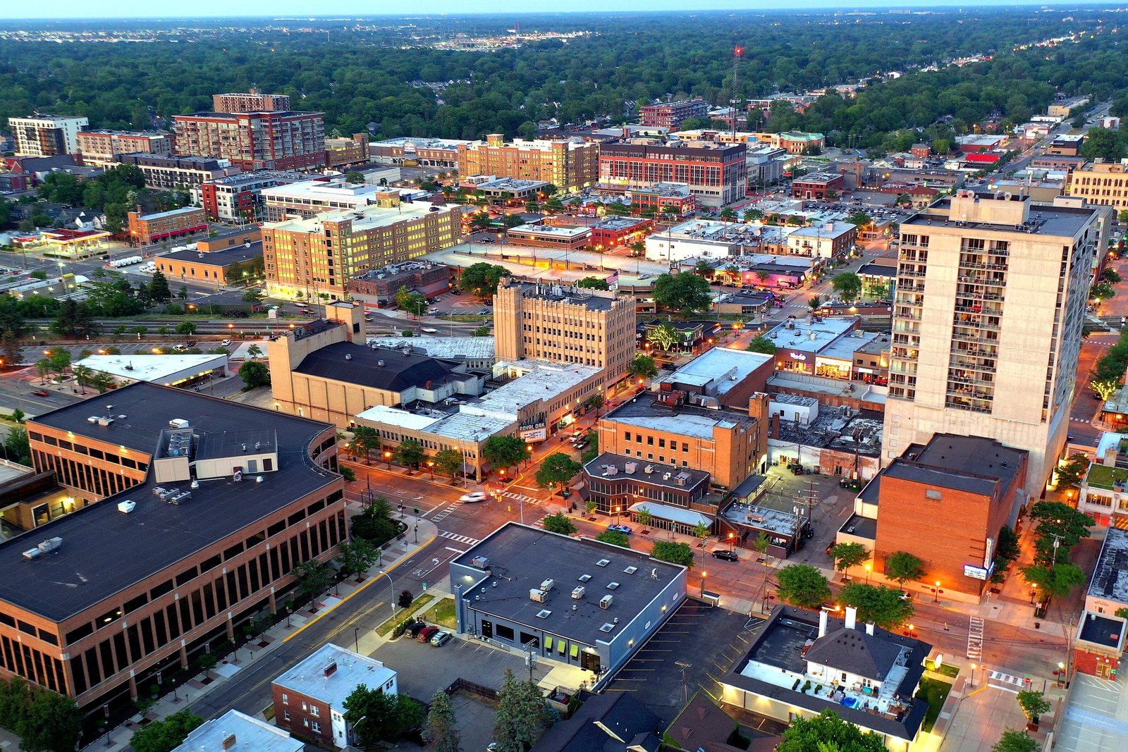 An aerial view of a downtown at dusk with brick buildings, a tall white tower, streets with cars and trees stretching to the horizon.