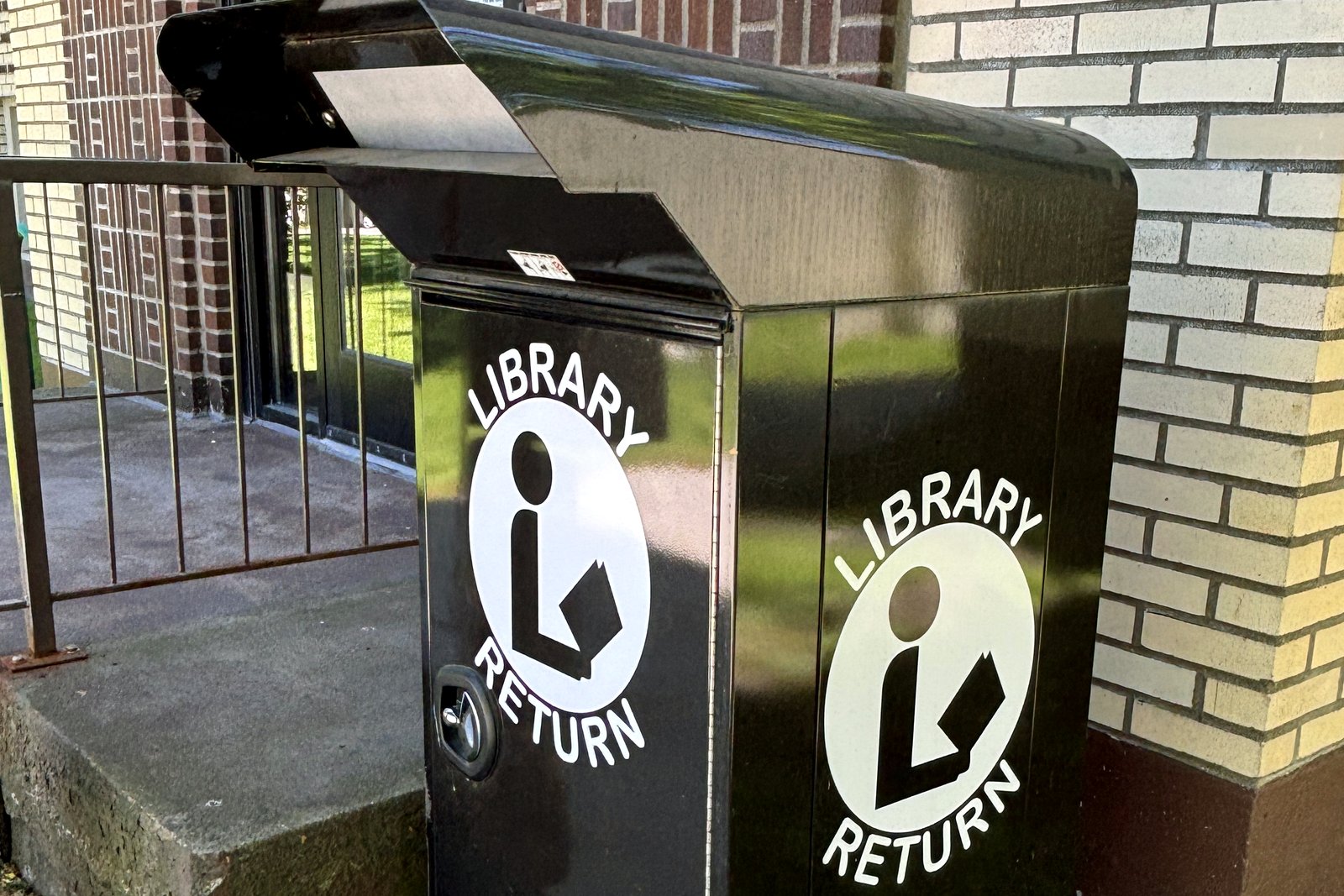 A black library book return bin outside a brick building, with a white book-return logo on its side.