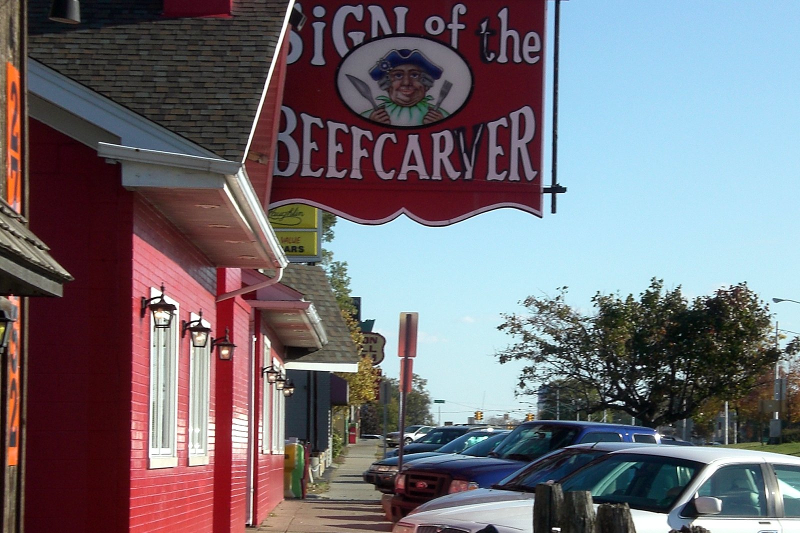 A hanging wooden sign with red lettering above the brick front of a restaurant along a sidewalk, with parked cars nearby.