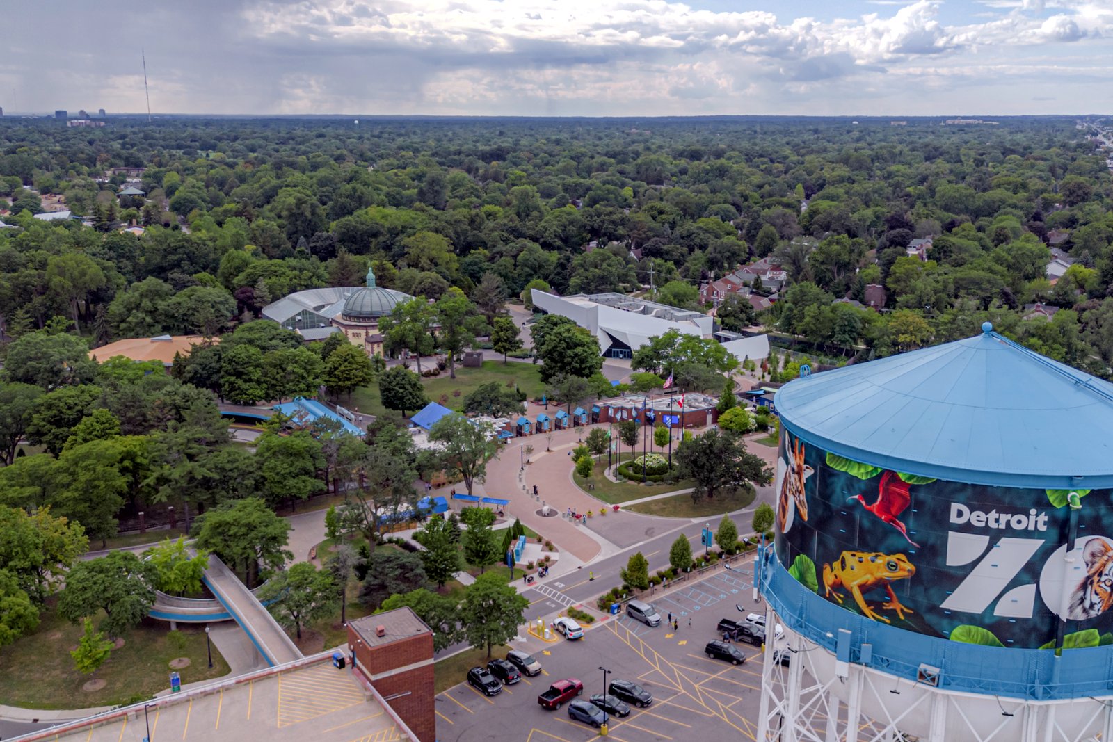 An aerial view of a zoo surrounded by trees, with paths, buildings, a parking lot and a tall water tower painted with the word Zoo.