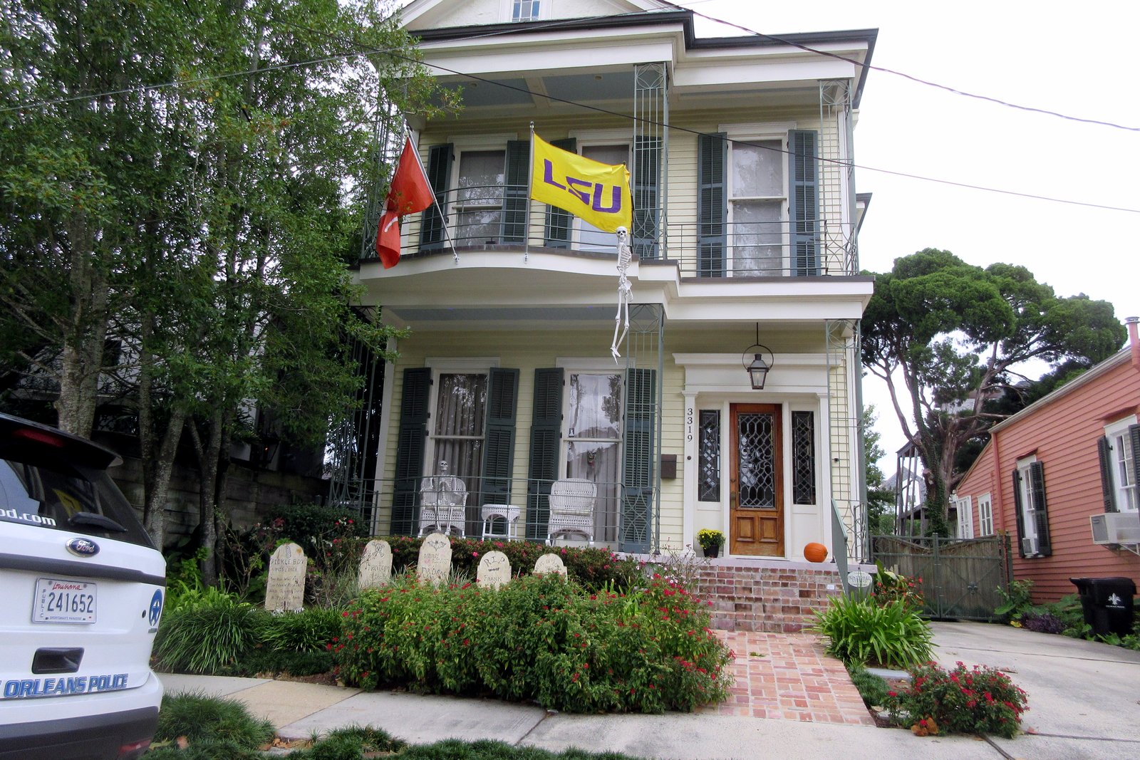 A two-story porch house with a yellow flag and a front-yard Halloween graveyard of small tombstones behind a low hedge.