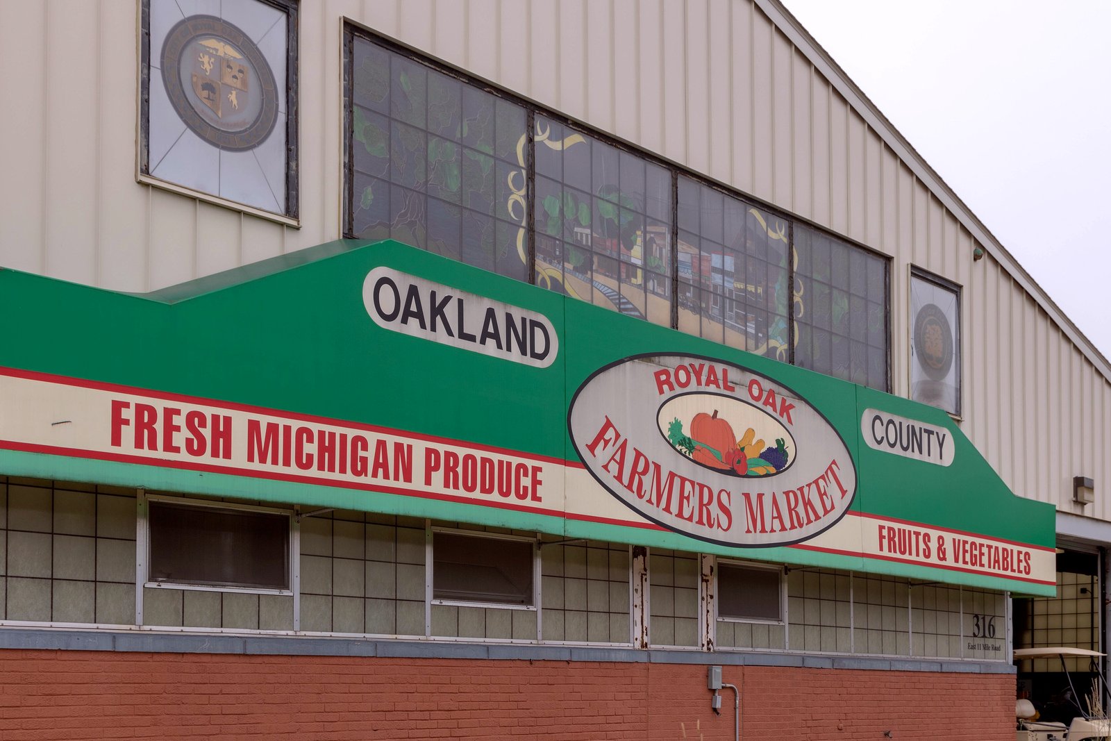 The front of a market building with a green awning reading Oakland County Fresh Michigan Produce Farmers Market, and a round sign above the entrance.