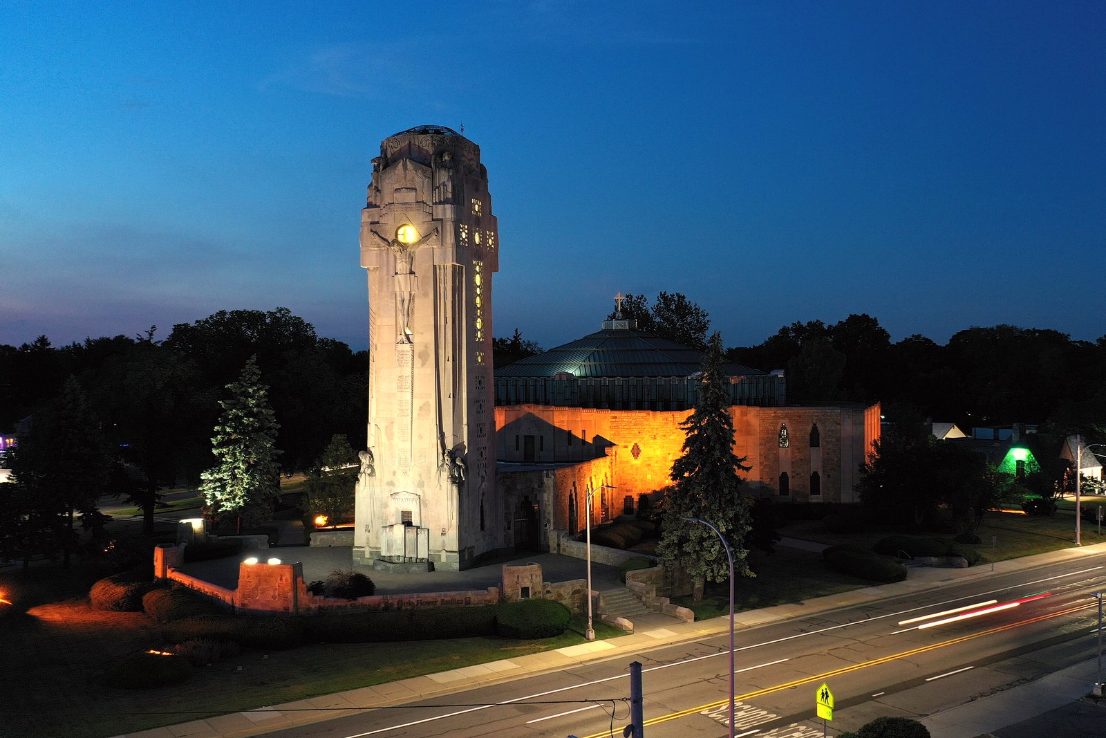 A tall stone Art Deco tower lit by floodlights at dusk, with a church building beside it and a street in the foreground.
