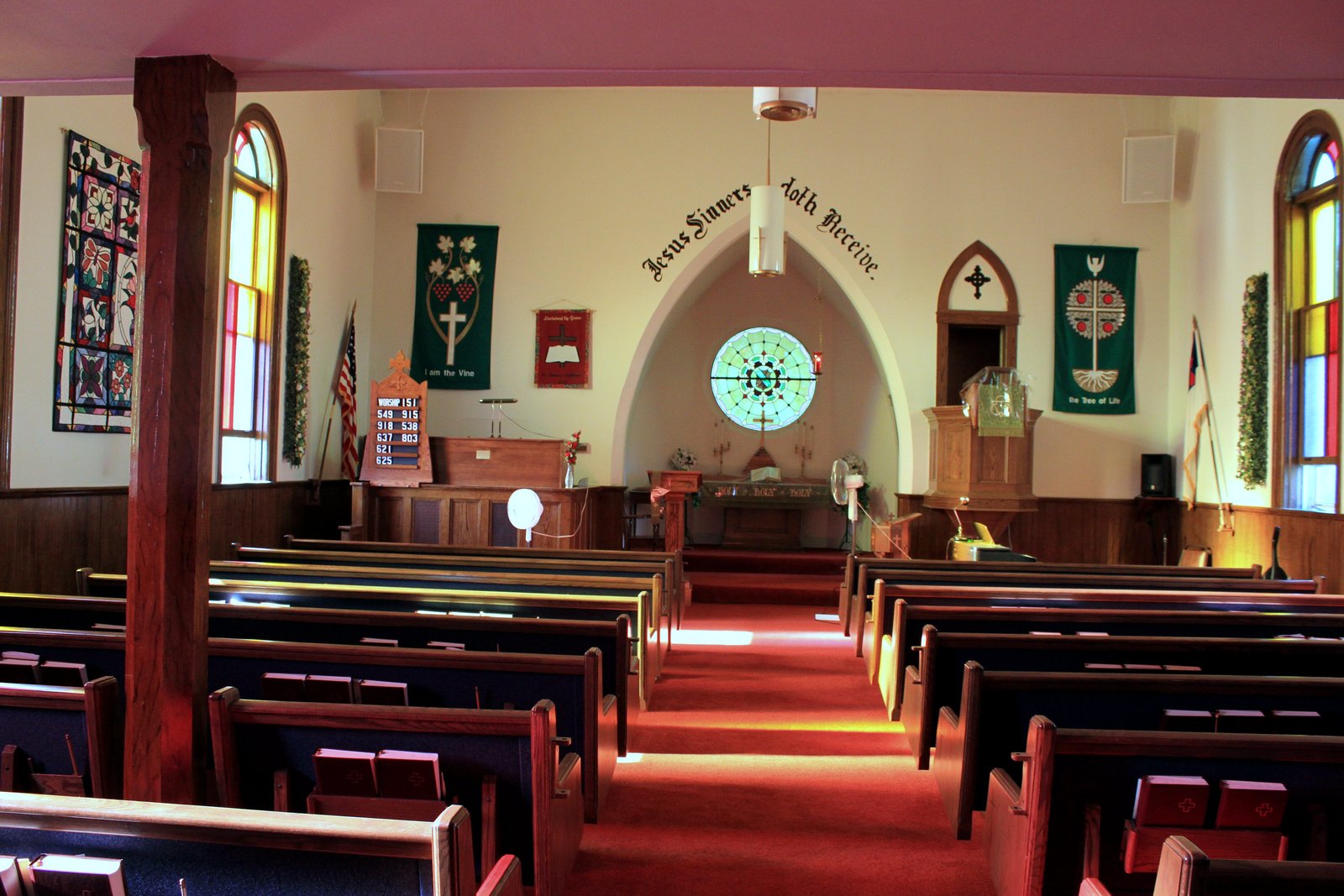 Rows of dark wooden pews with red cushions face a carpeted aisle leading to an altar under an arch, with a round stained-glass window and green banners on cream walls.