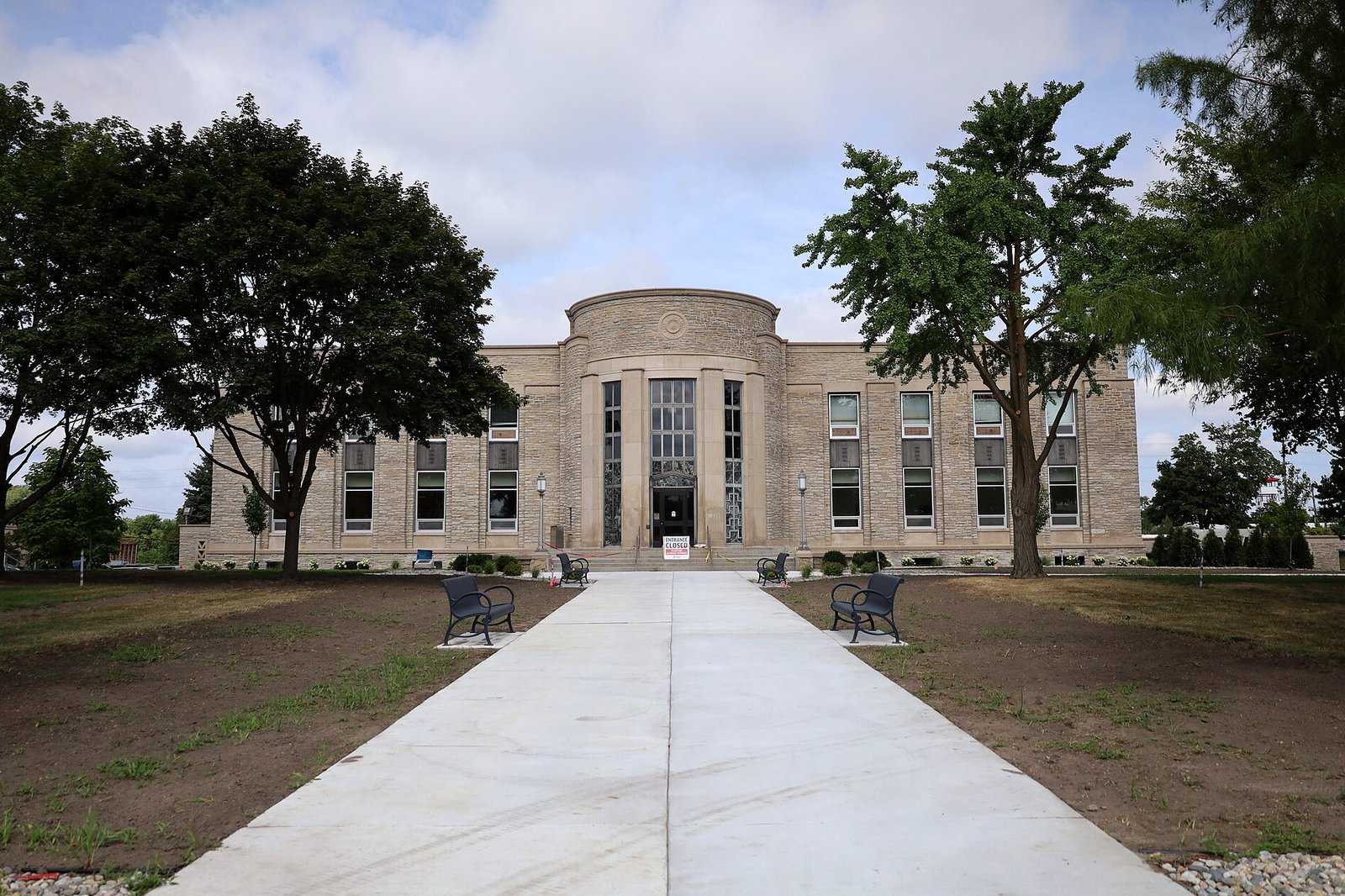 A pale stone civic building with a rounded central entrance tower and tall windows, reached by a long concrete walk between young lawns and leafy trees under a cloudy sky.