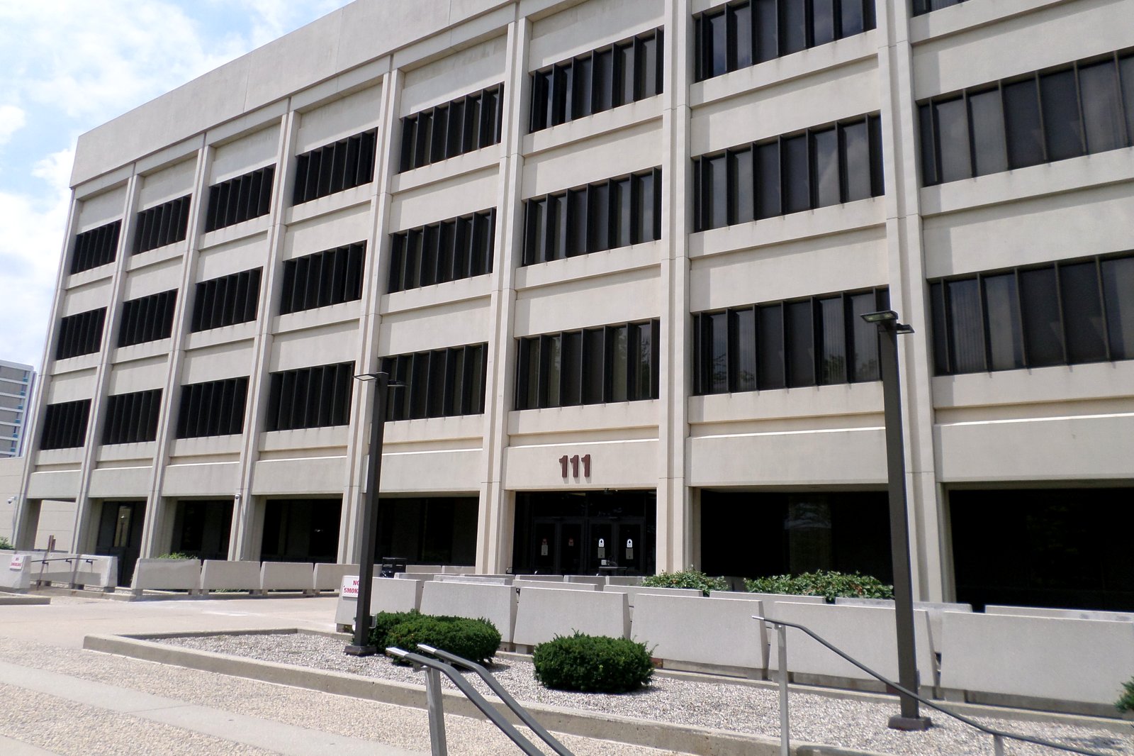 A tall, light-colored office building with rows of dark windows and a small plaza and planters in front, under a partly cloudy sky.