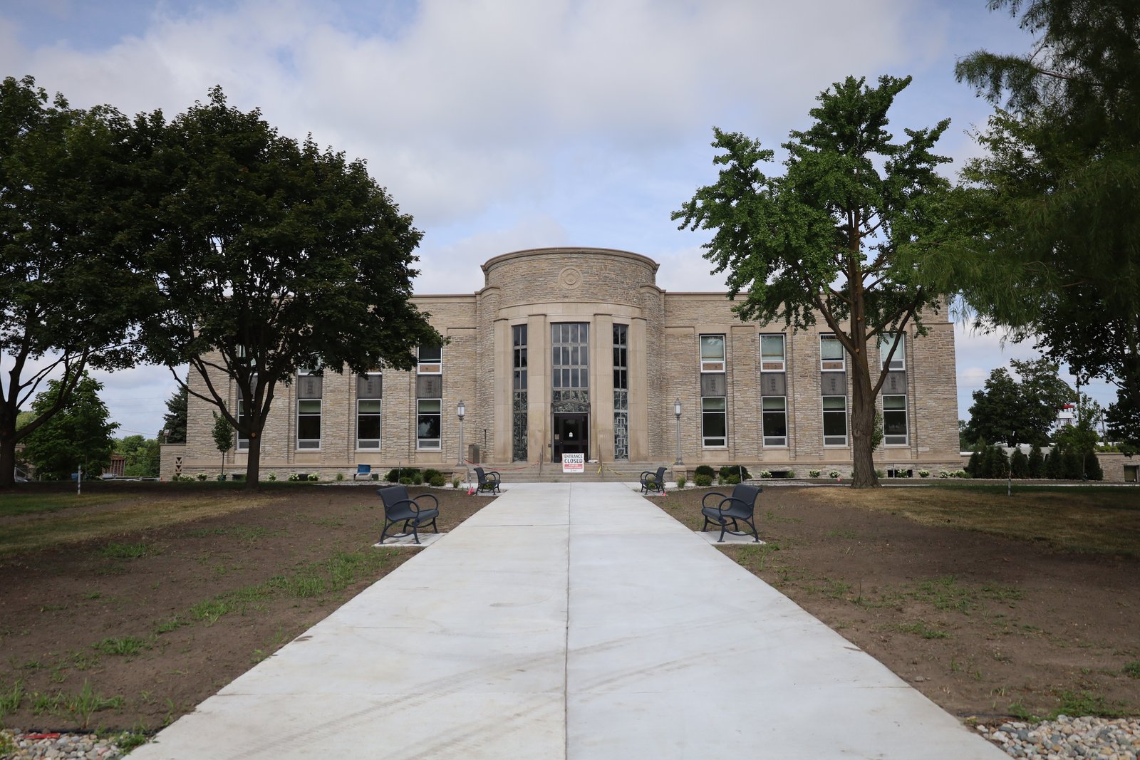 A two-story limestone city hall with a rounded central entrance and tall windows, reached by a new concrete walkway lined with benches and trees.