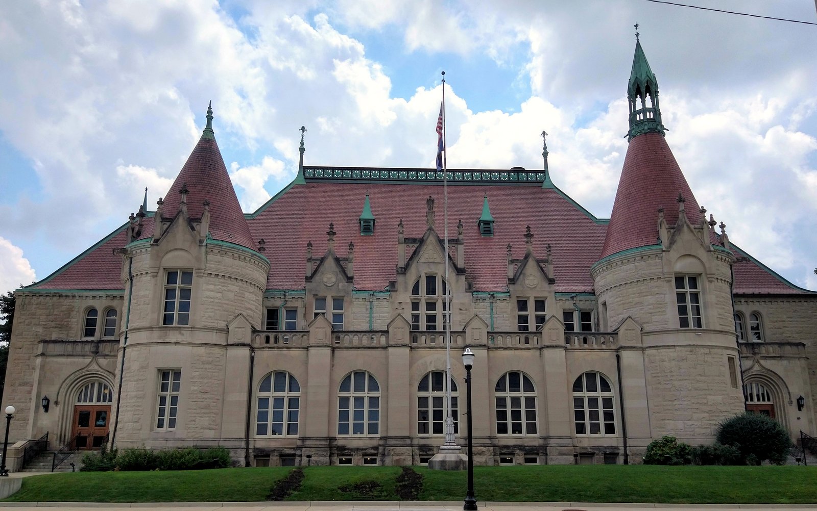 The Castle Museum of Saginaw County History, a tan stone building with red-roofed turrets and a central gable, stands behind a green lawn under a partly cloudy sky.
