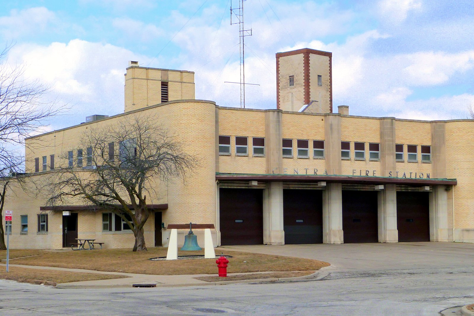 A tan brick fire station with four brown bay doors, a square tower and a bell on a stand out front, under a partly cloudy sky.