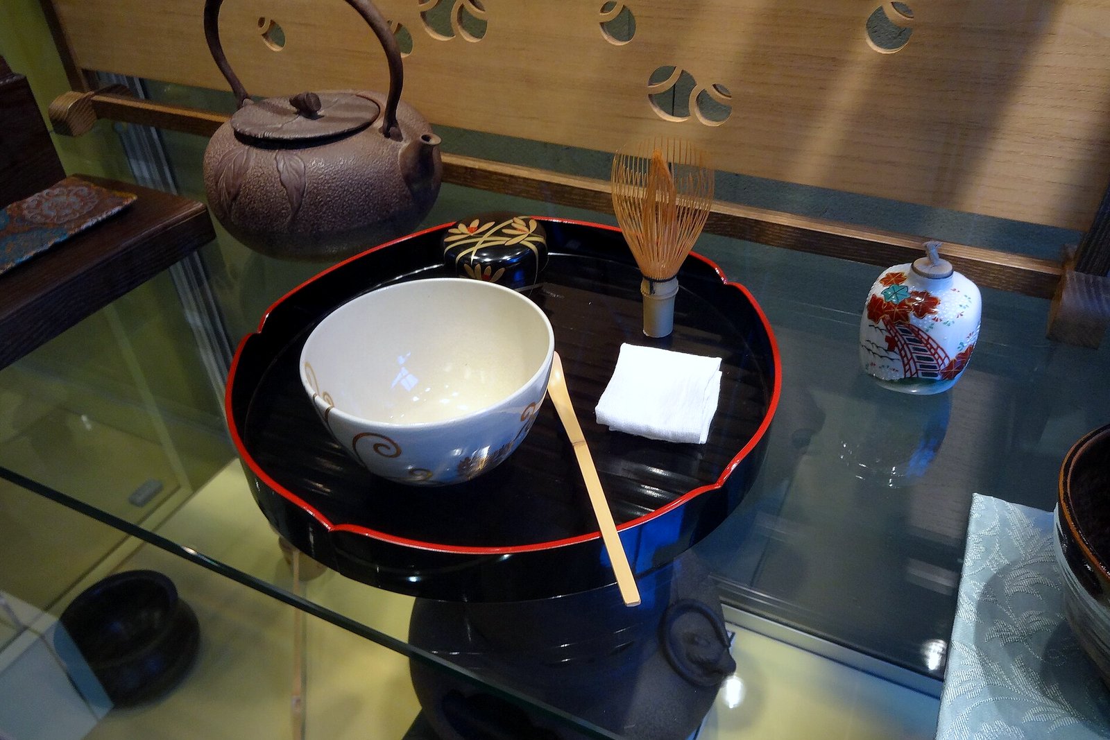 A lacquered tray holding a white tea bowl, a bamboo whisk and a bamboo scoop, with an iron kettle and a small tea caddy, on display behind glass.