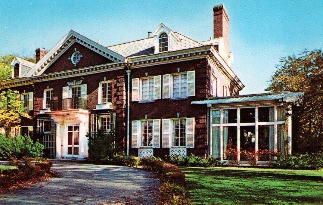 A large red-brick Georgian-style house with white shutters, a white columned entrance and a glass sunroom wing, set behind a curving drive and green lawn.