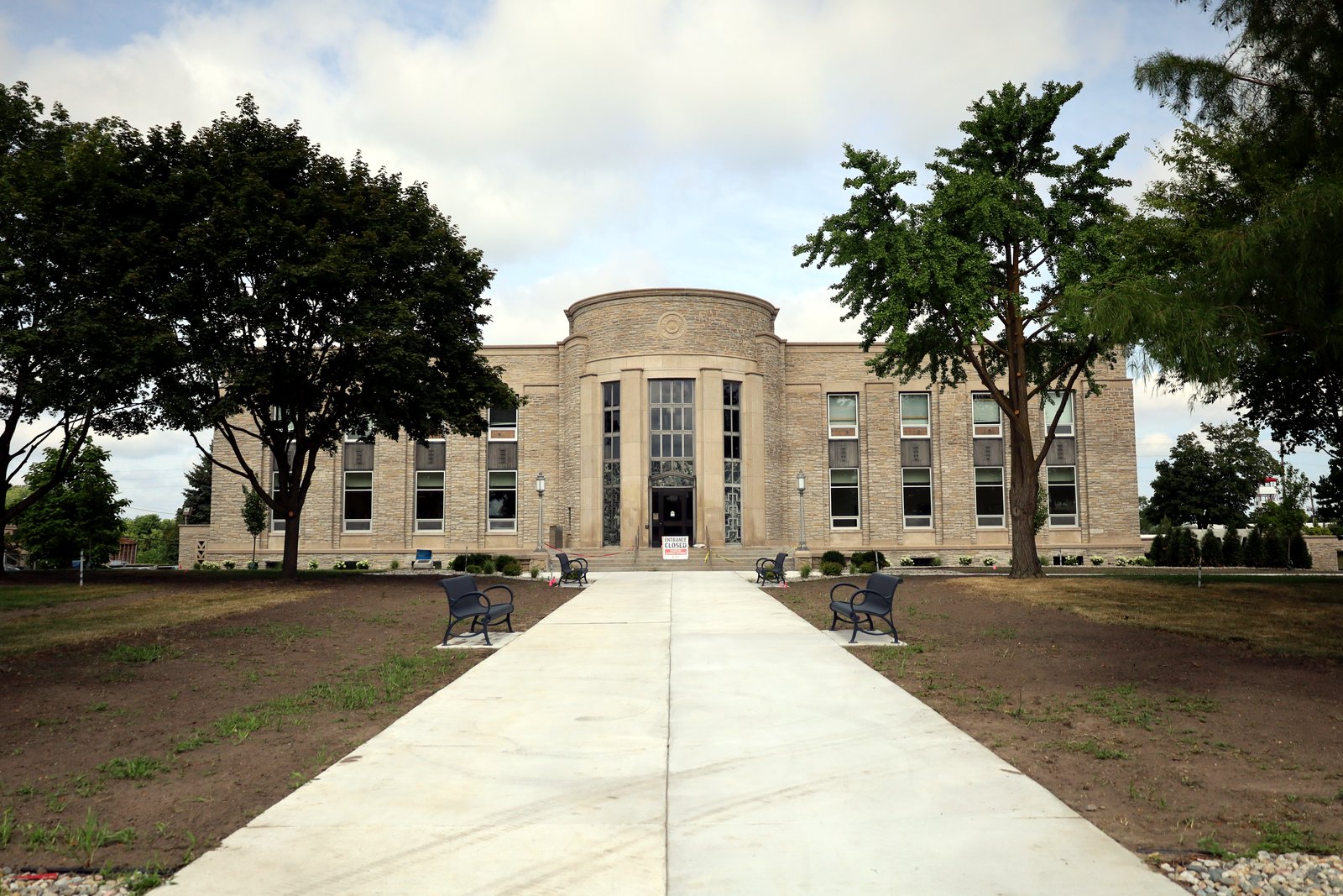 A tan stone civic building with a rounded central entrance tower, flanked by trees, at the end of a long concrete walkway with benches.