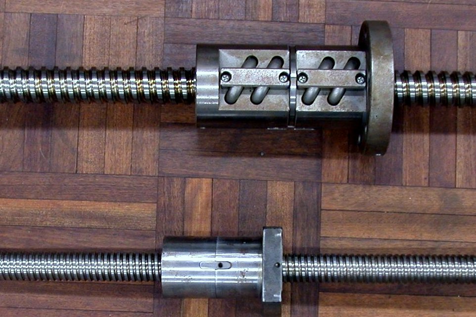 Two long steel ball screws with threaded shafts and nut assemblies lying on a parquet floor.