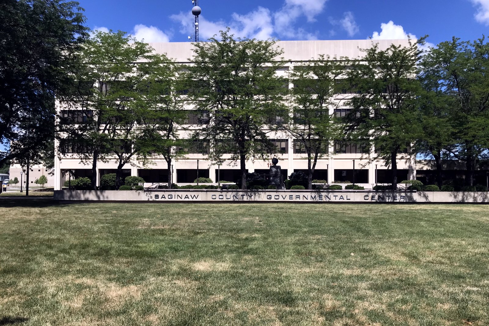 A long, four-story concrete government building behind a row of trees, with a low wall in front reading Saginaw County Governmental Center and a wide lawn in the foreground.