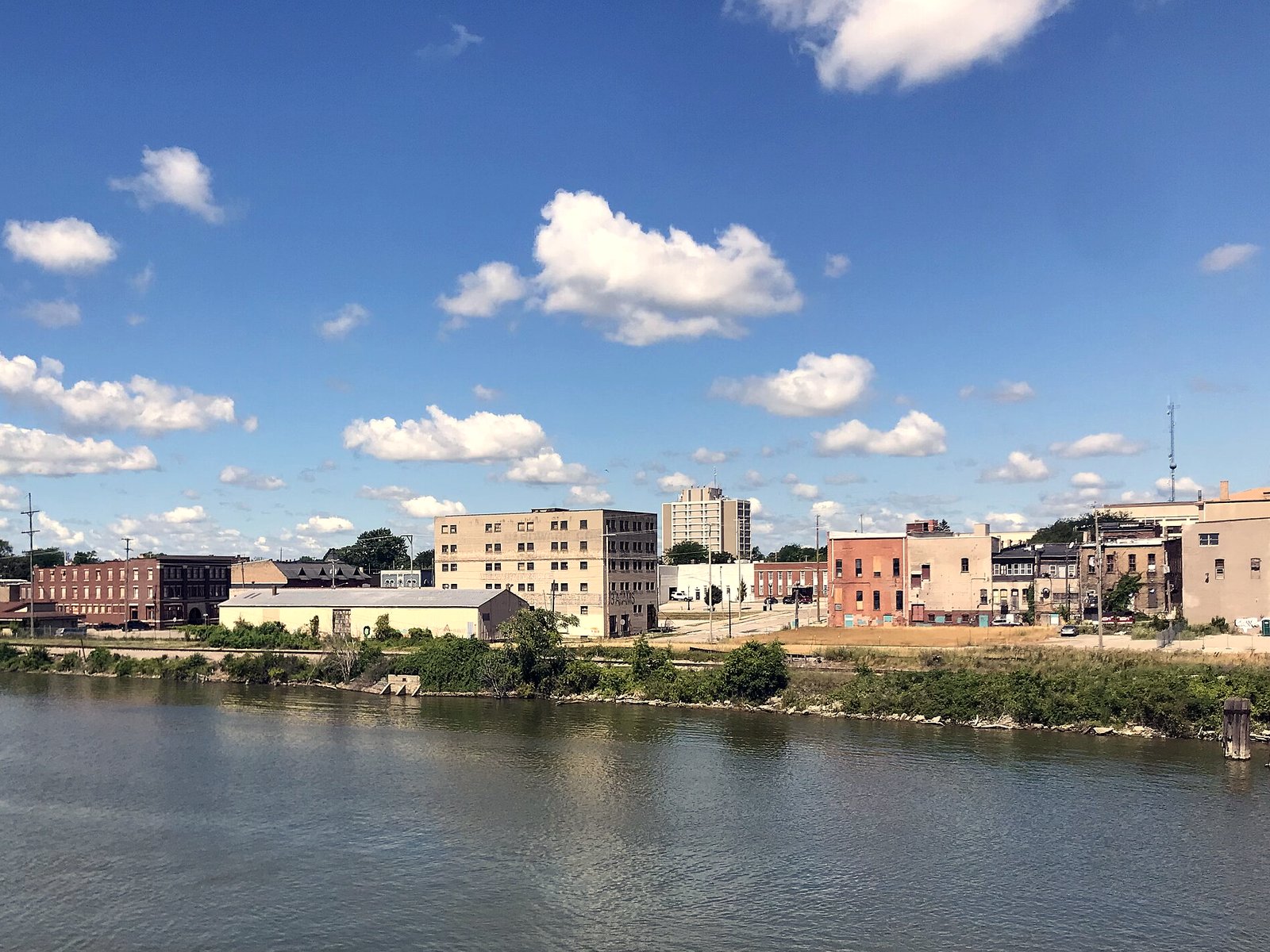 The west bank of the Saginaw River in Old Town Saginaw, with low brick commercial buildings, a pale six-story building and a taller tower behind a grassy riverbank under a bright blue sky with white clouds.
