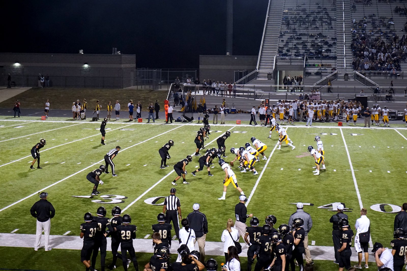 High school football players lined up at the line of scrimmage during a night game, with fans in the stands behind them.