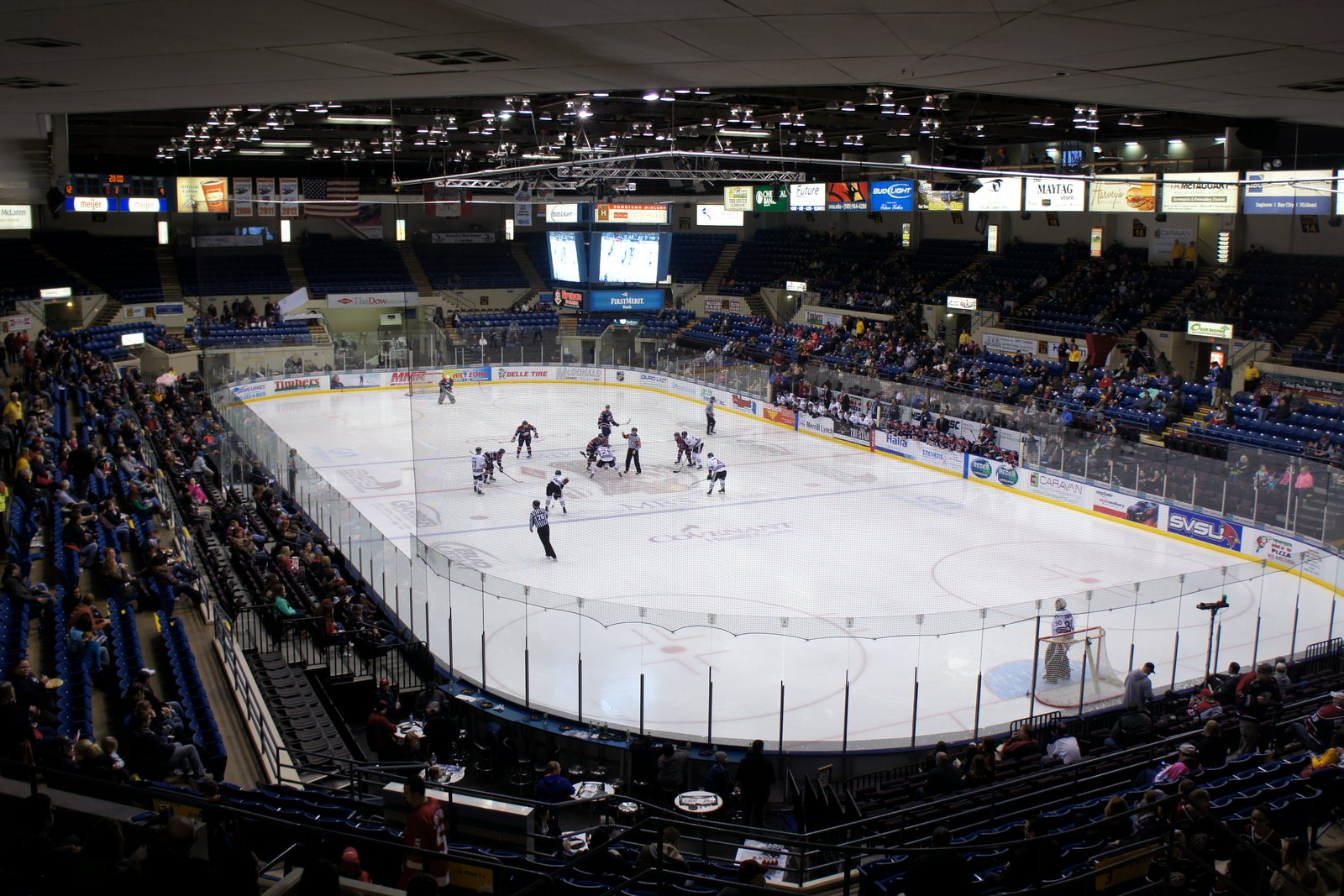 A hockey game in progress on a white rink inside an arena, seen from high in the stands, with fans in blue seats.