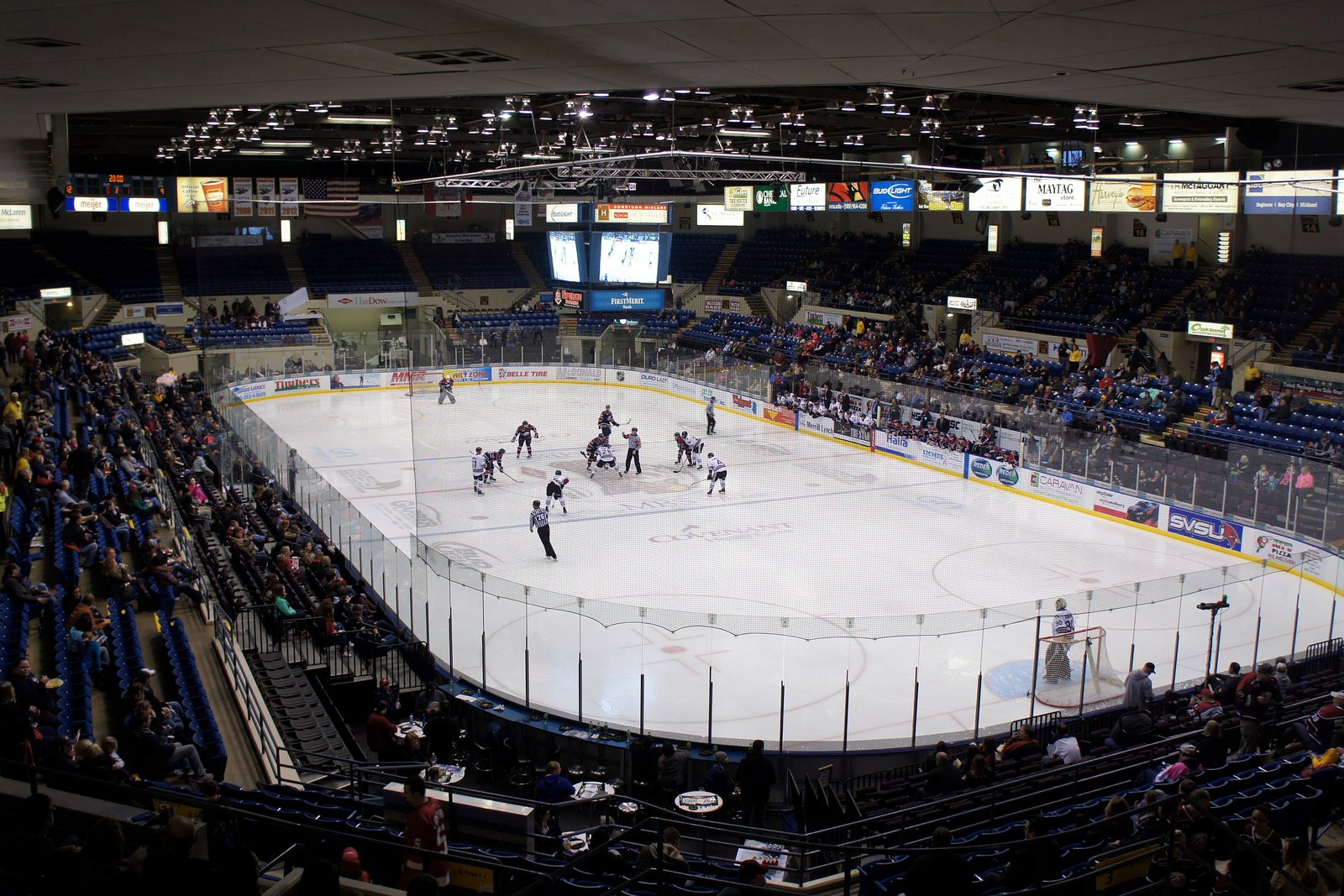 A hockey game in progress on a white rink seen from high in the stands of an indoor arena, with fans in the lower seats and lit advertising boards along the upper level.