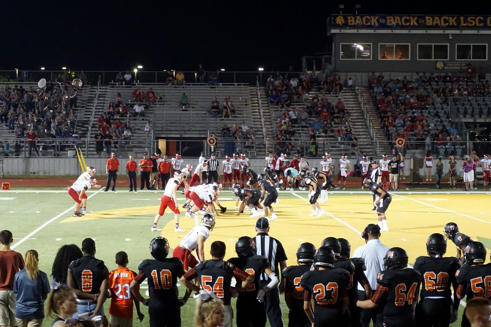 High school football players in orange and black jerseys watch from the sideline as a play unfolds at night.