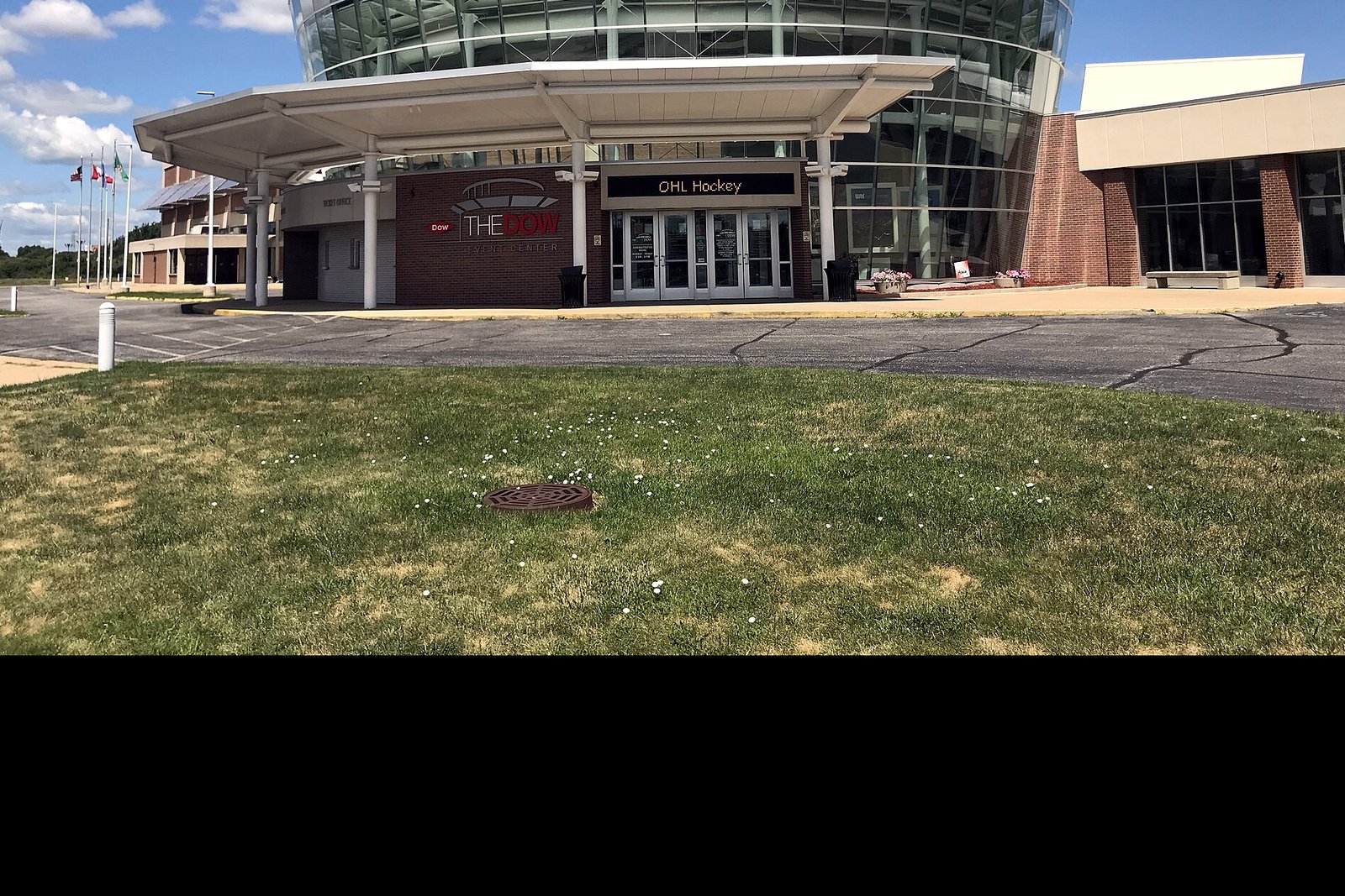 The glass-fronted Dow Event Center building with a paved plaza and landscaping in front under a partly cloudy sky.
