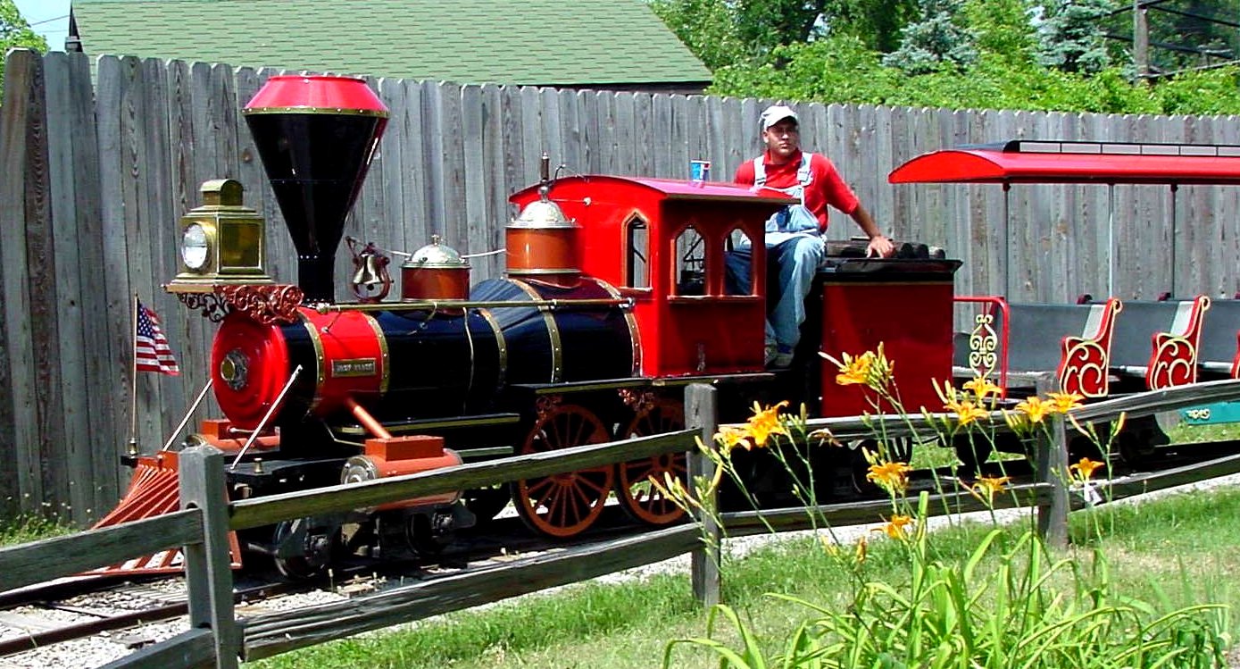 A small red and black steam-style train with a conductor riding along a fenced track beside yellow flowers.