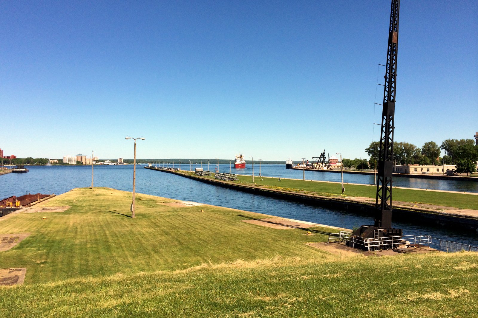 A grassy point and a long concrete pier beside blue water under a clear sky, with a freighter in the distance heading toward the locks.
