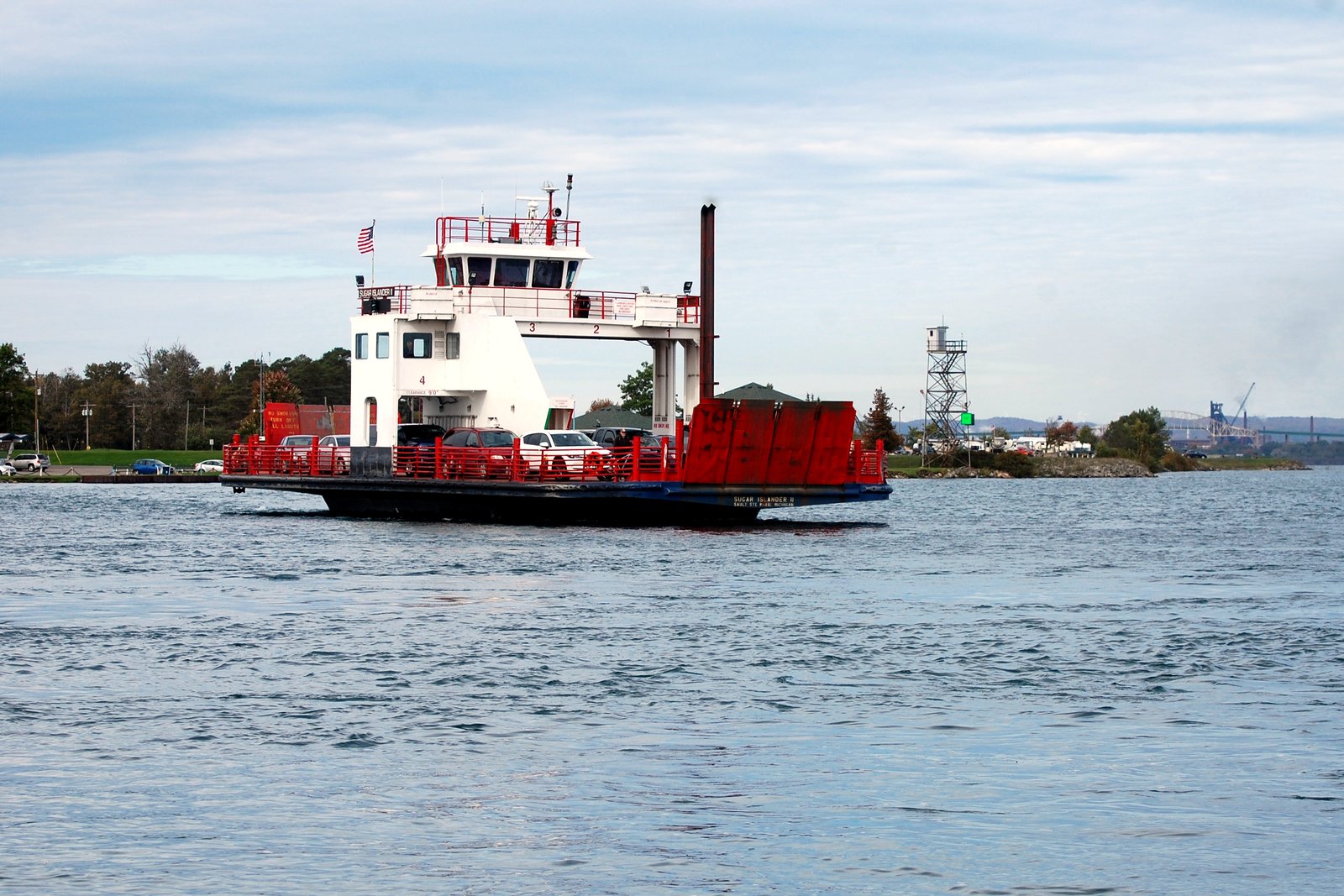 A red-and-white car ferry with cars on its open deck crosses a river, with a low shoreline behind it.