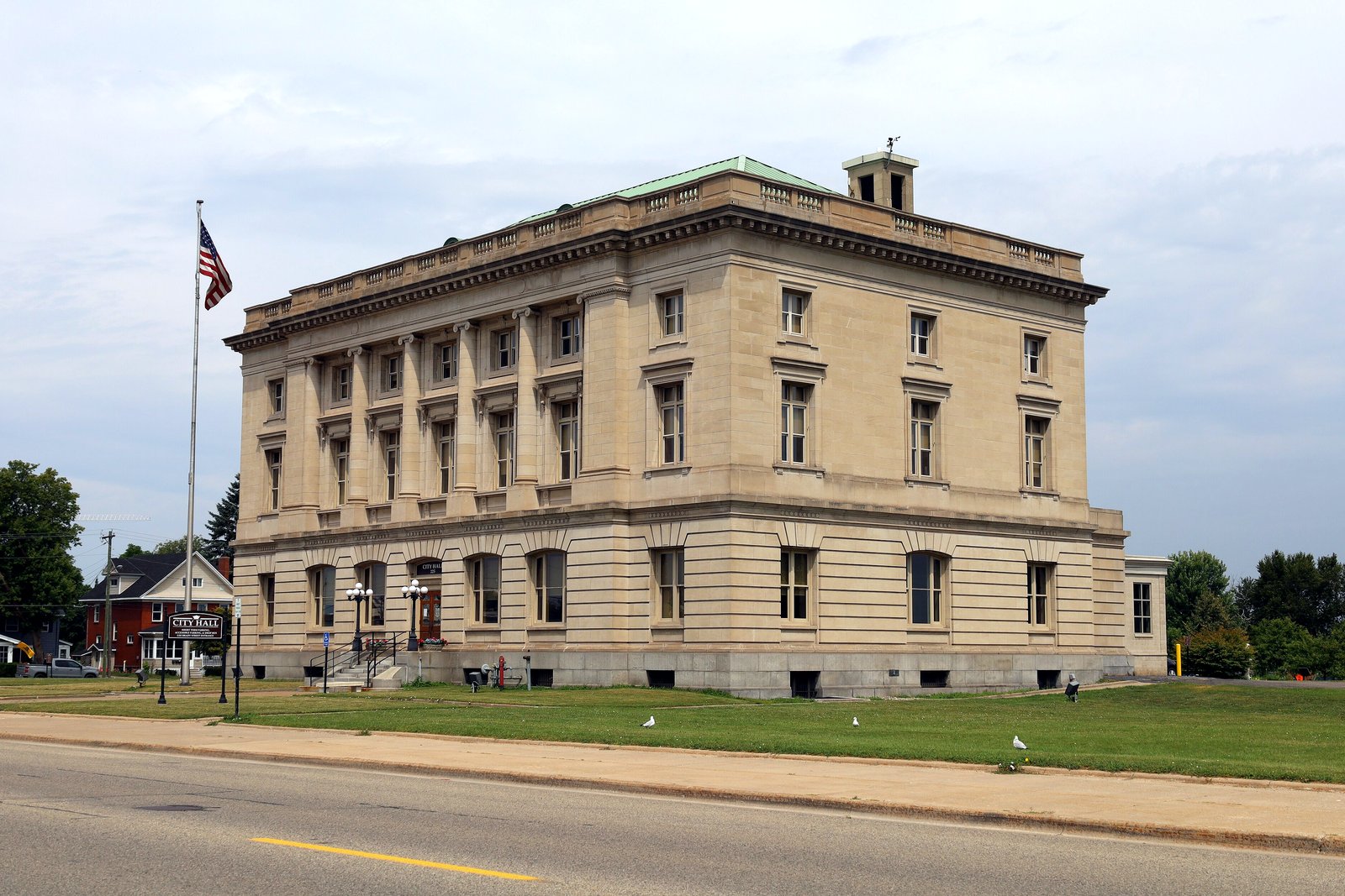 The front of Sault Ste. Marie City Hall, a stone building with a flagpole, seen across a street.