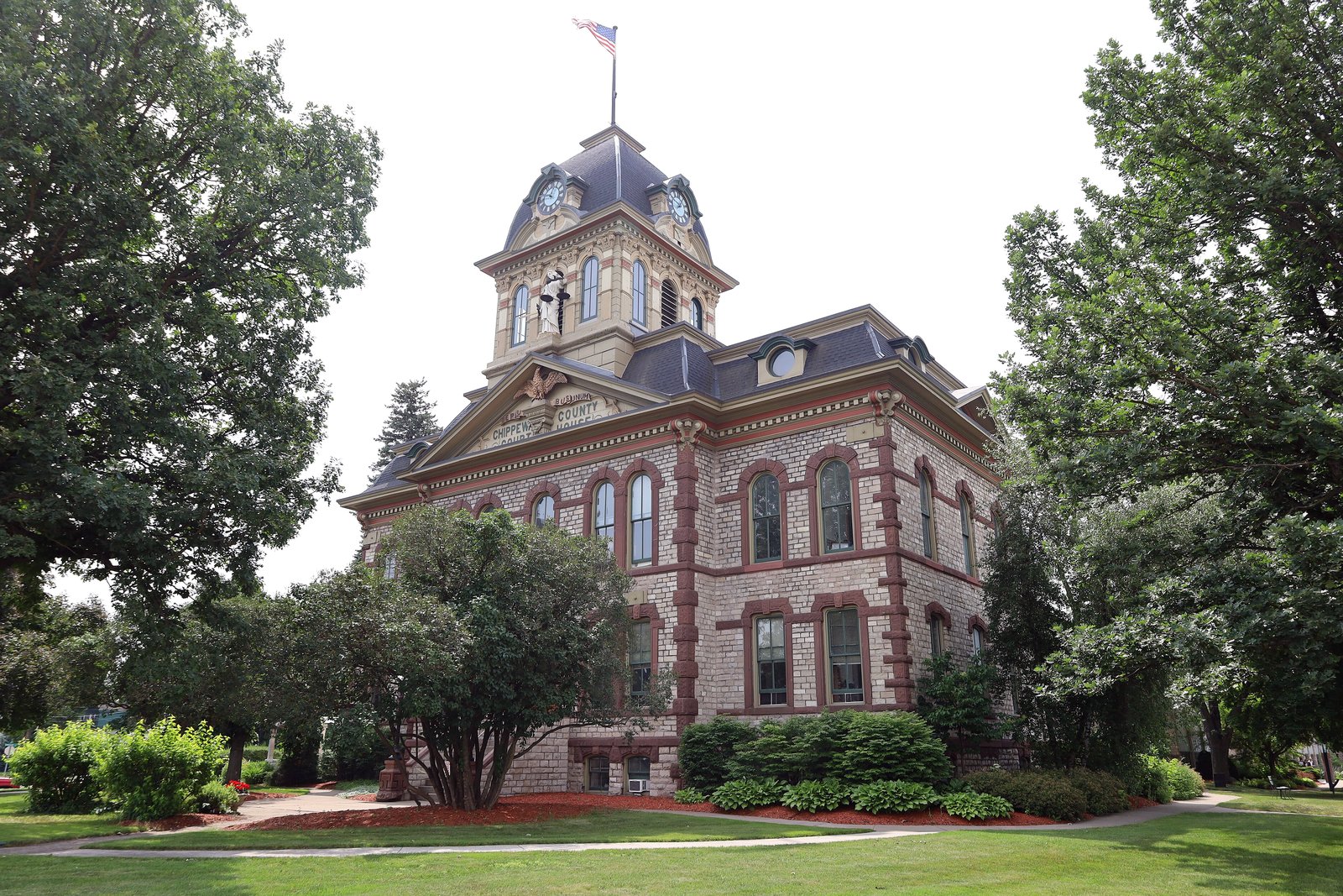 A red brick 19th-century courthouse with a clock tower and cupola behind trees and a manicured lawn.