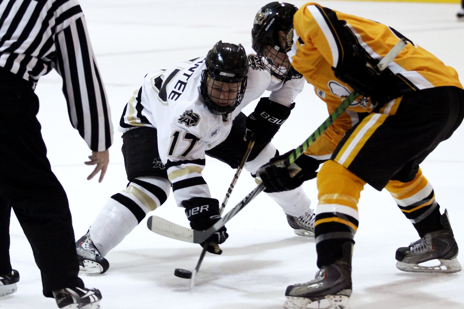 A hockey player in a white jersey wins a faceoff against a player in black and gold as a referee watches.